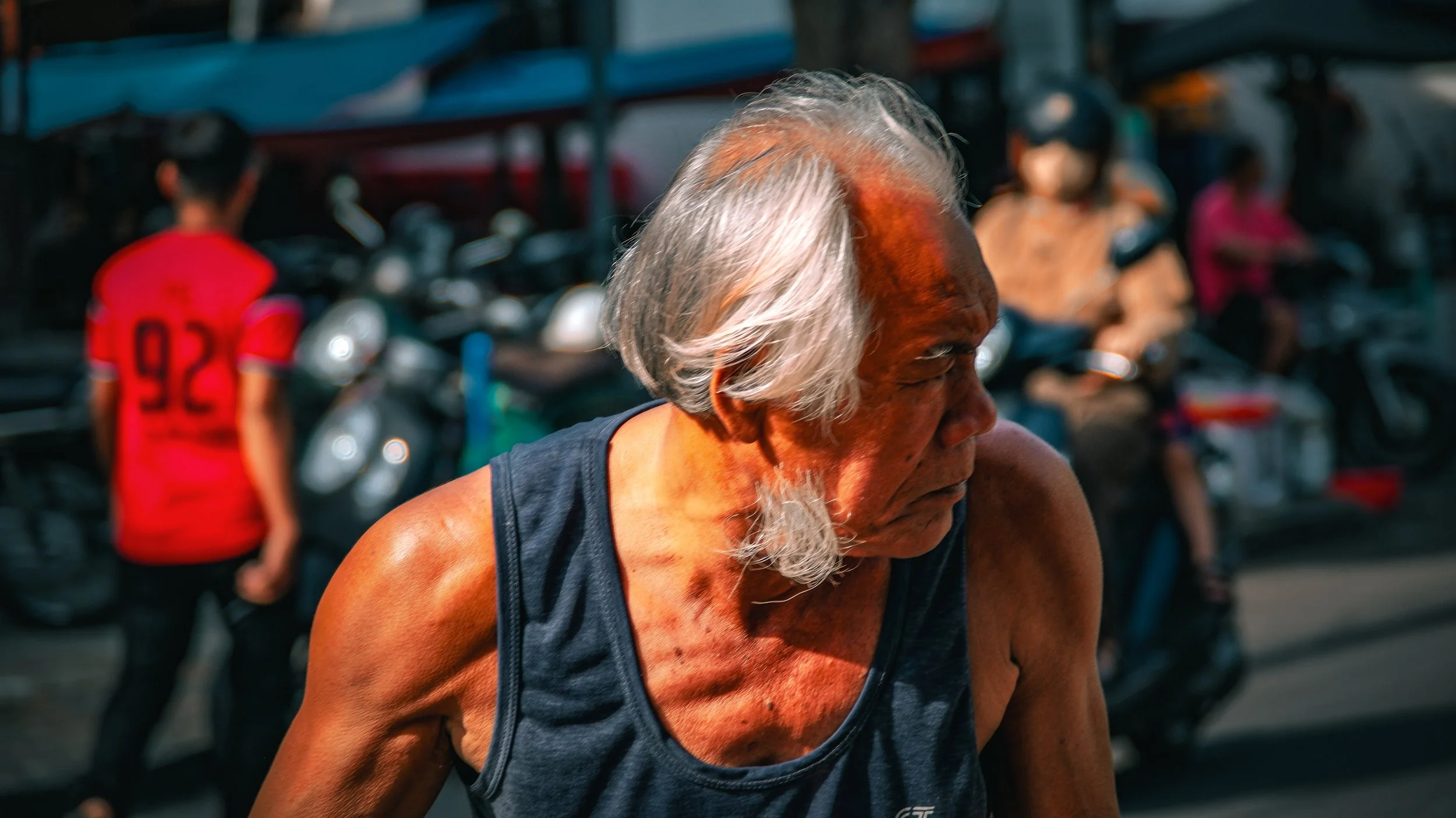 A senior man with gray hair and a black sleeveless shirt appears to be exercising outdoors among motorcycles and other people.
