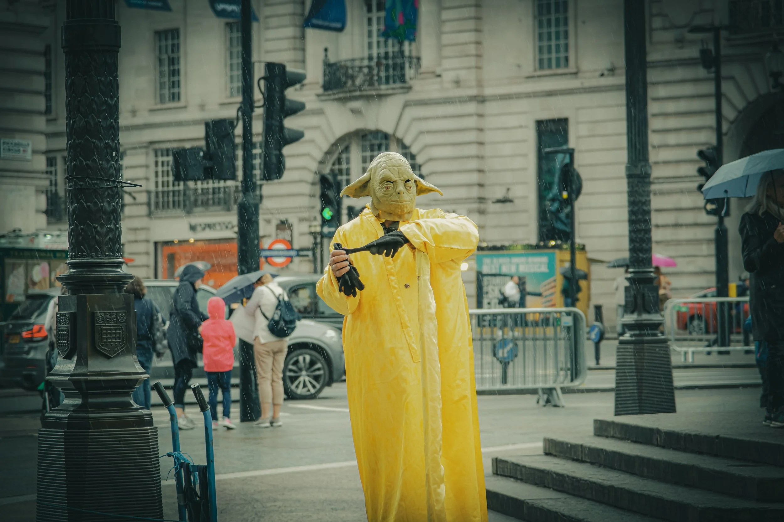 Person dressed as Yoda from Star Wars, wearing a yellow raincoat and rain gloves, standing in a rainy urban street.