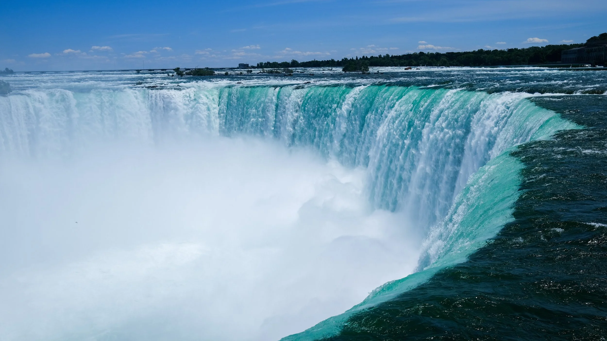 The image shows the Niagara Falls with water cascading over the edge, mist rising, and a partly cloudy sky above.