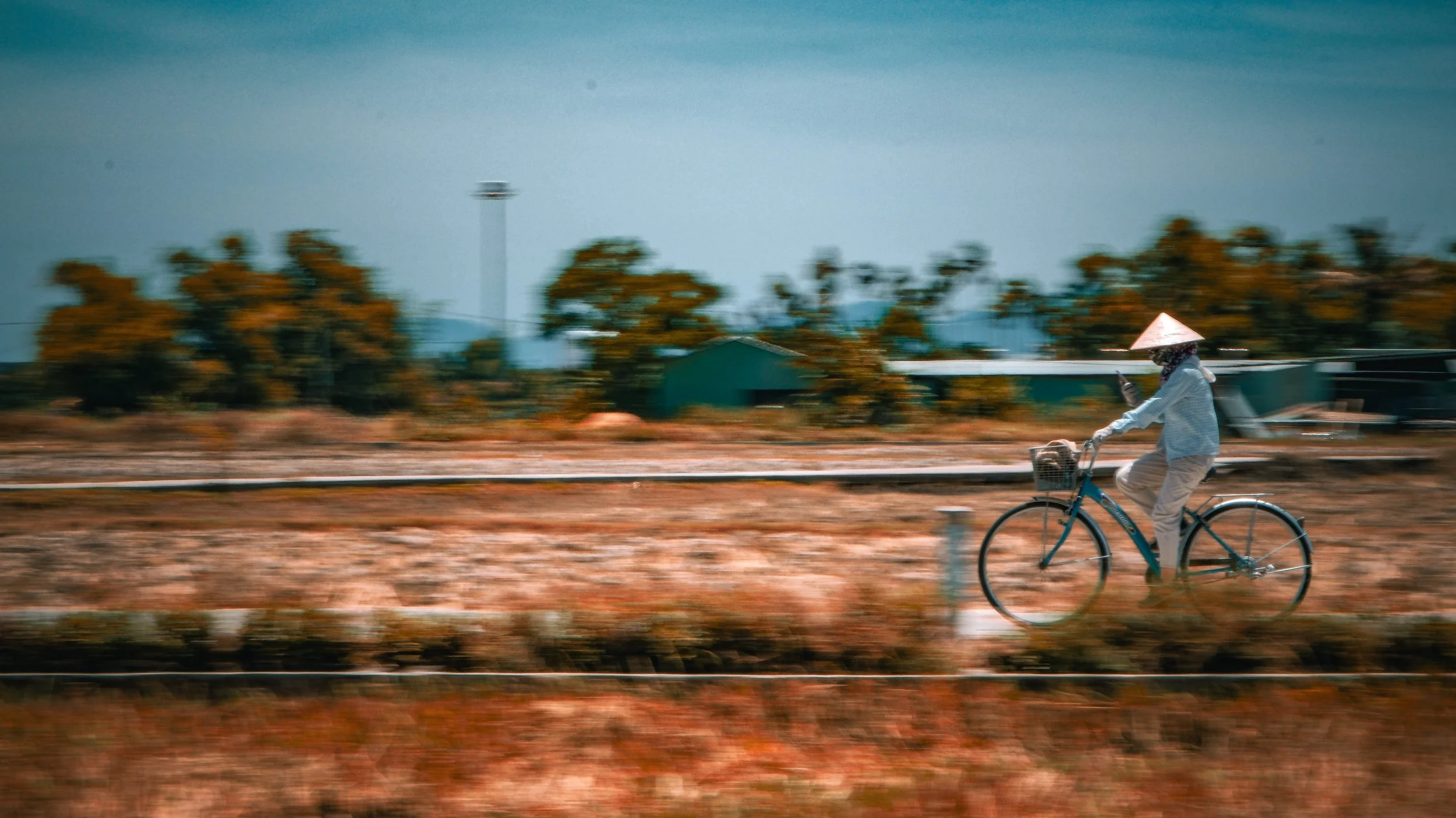 A person riding a bicycle on a rural road, wearing a conical hat and light clothing, with trees and buildings in the background under a clear sky.
