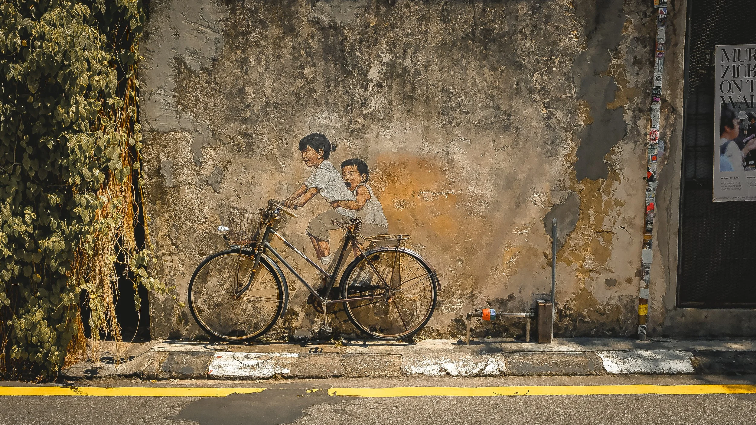 A mural of two children riding a bicycle, painted on a weathered wall with peeling paint. The scene shows an older child pedaling and a younger child sitting on the back, looking happy. There is green foliage on the left side of the mural and a bicyc