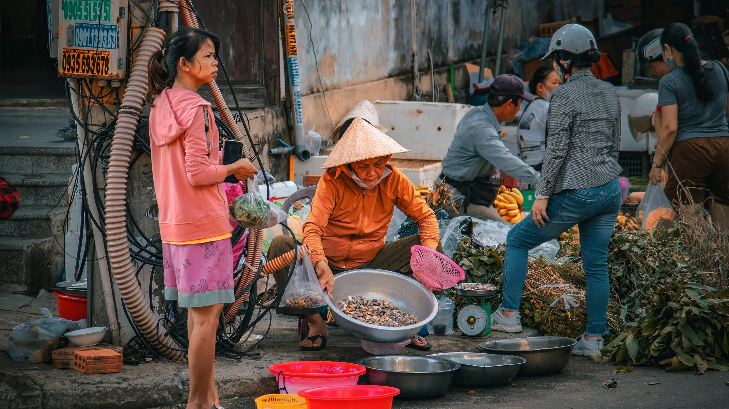 Street market scene with women selling clams and vegetables. One woman wears a traditional conical hat, while others are engaged in buying and selling. The background shows a worn wall, electrical wires, and various market supplies.