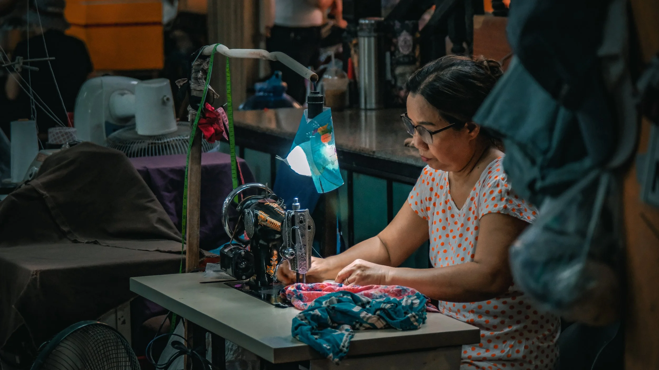 A woman sewing fabric at a worktable inside a workshop, with sewing tools, fabrics, and equipment around her.