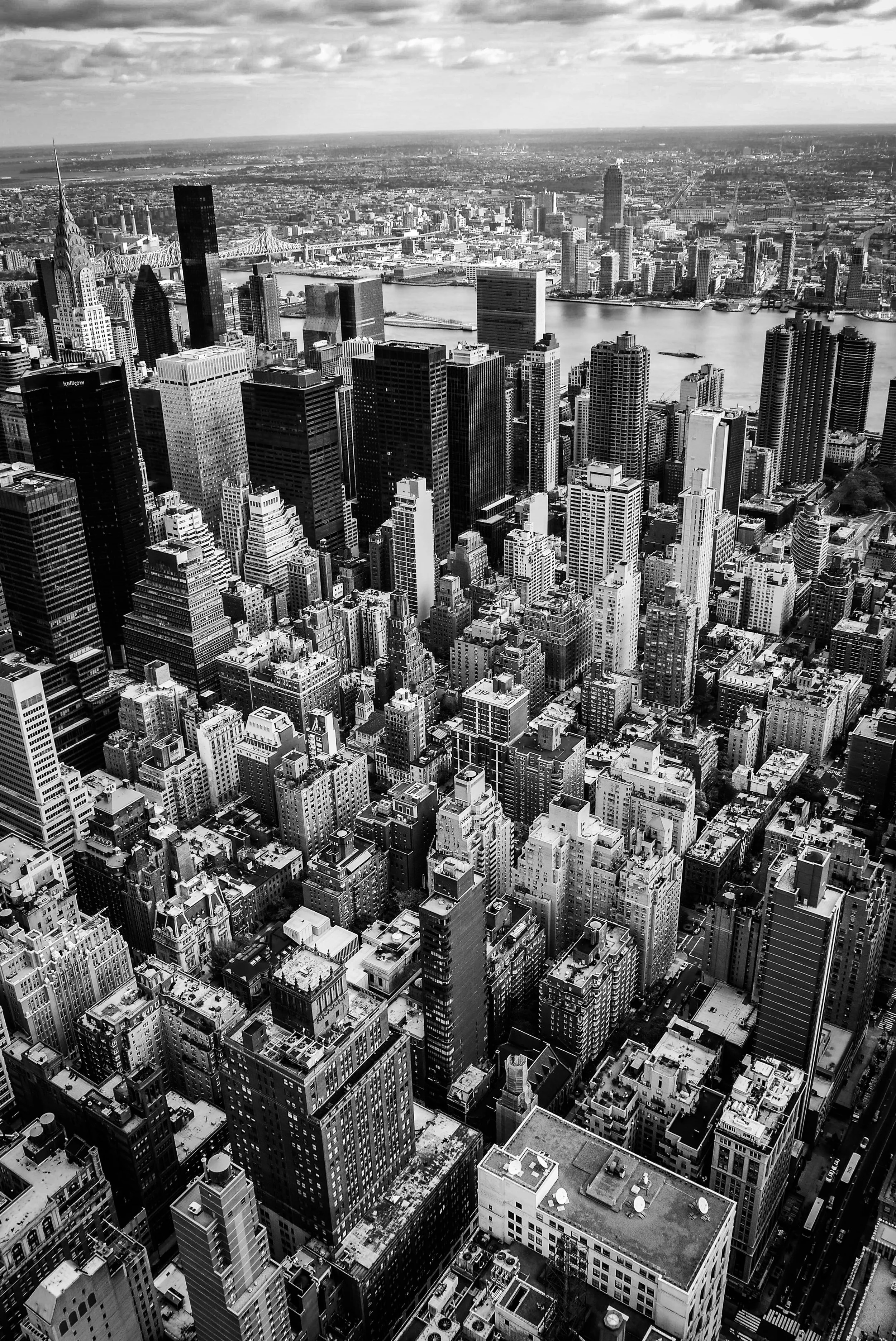 Black and white aerial view of New York City skyline showing numerous skyscrapers, the East River, and a bridge, with a cloudy sky overhead.