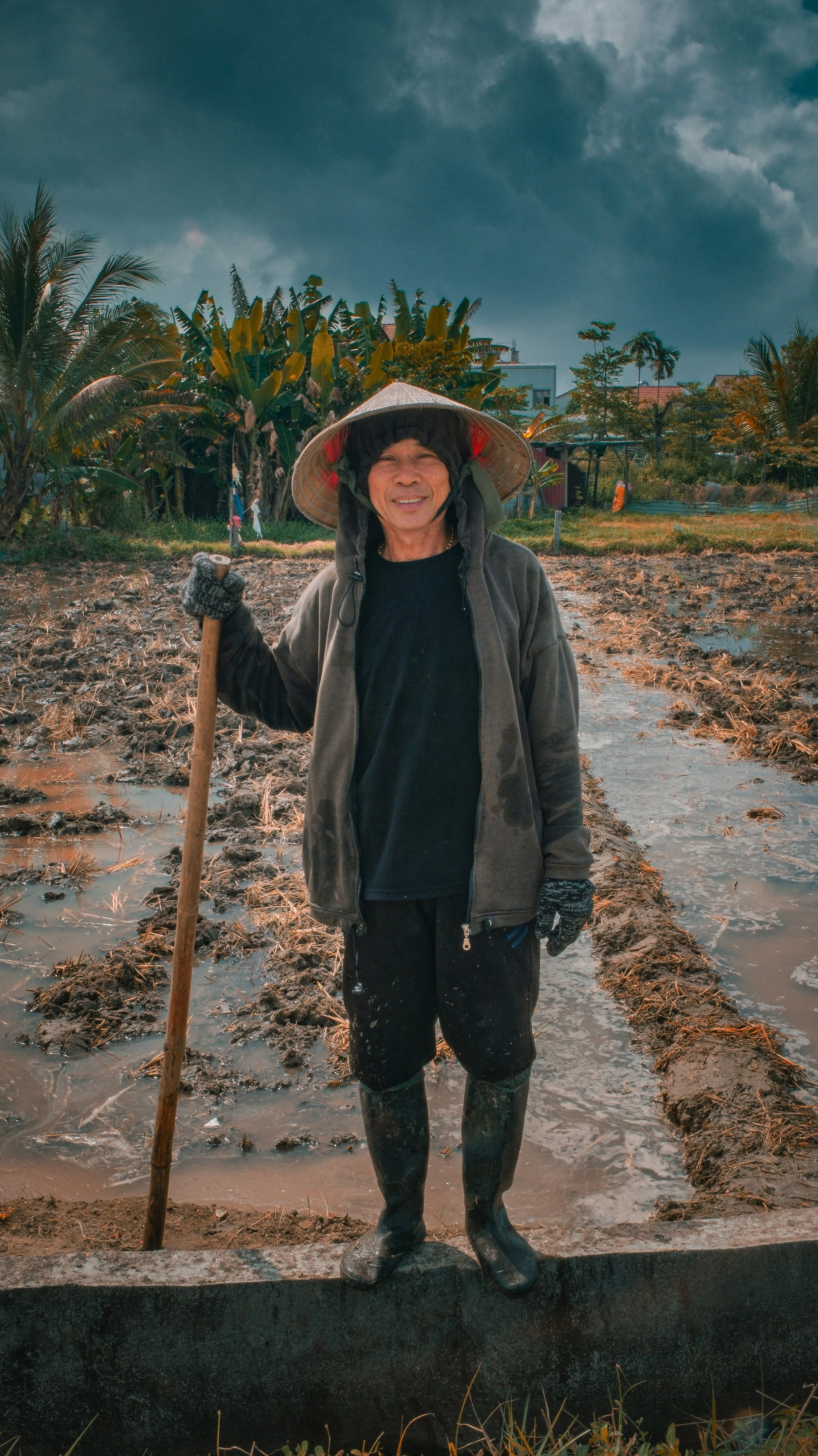 A man wearing a conical hat, hoodie, gloves, and boots, standing in a muddy field holding a hoe, with a cloudy sky and tropical trees in the background.