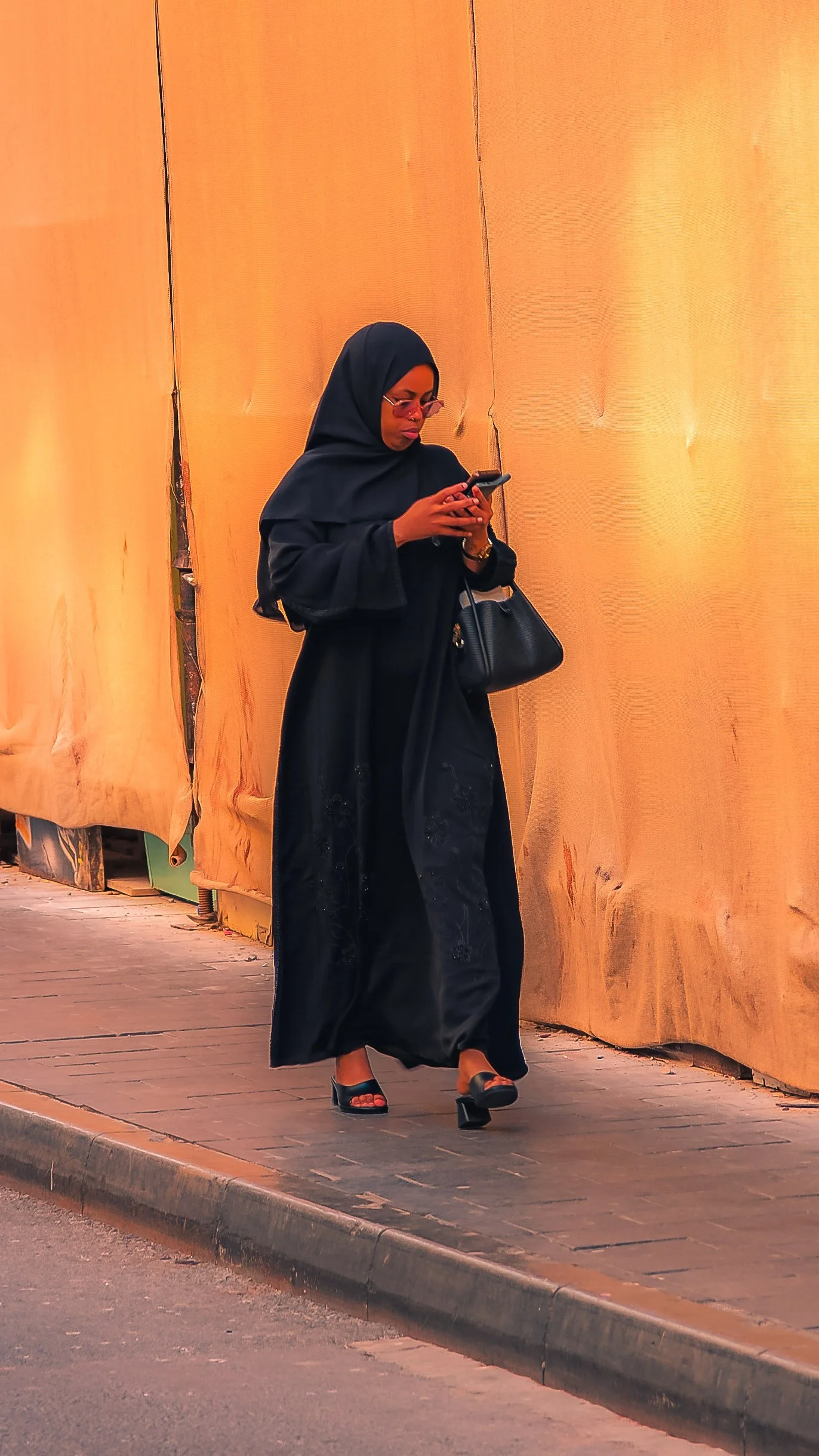 A woman dressed in black, wearing a headscarf, sunglasses, and open-toed heels, walking on a sidewalk against a large yellow-orange wall.