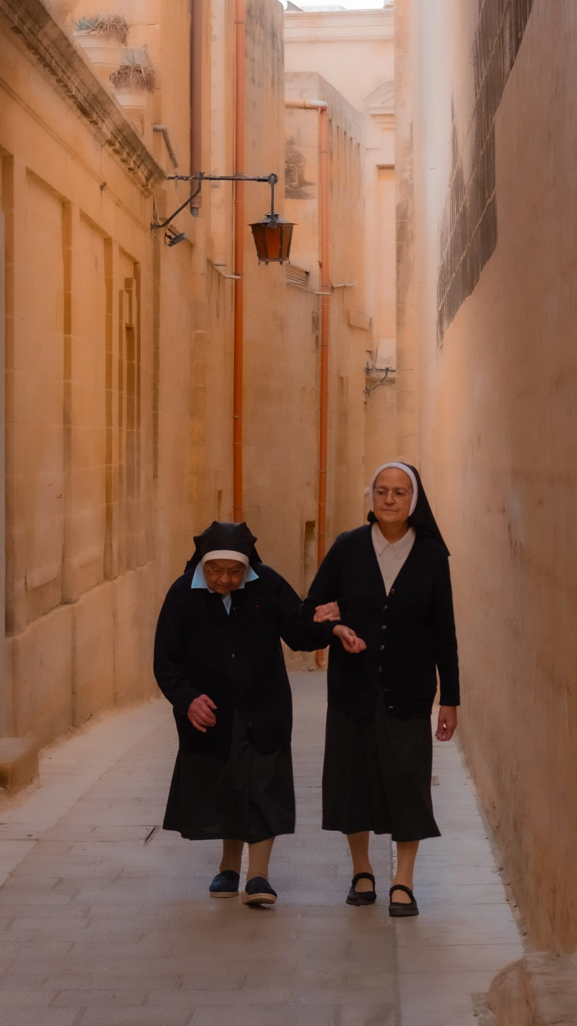 Two nuns walking arm in arm through a narrow stone alleyway with beige walls and a wrought iron lantern hanging from above.