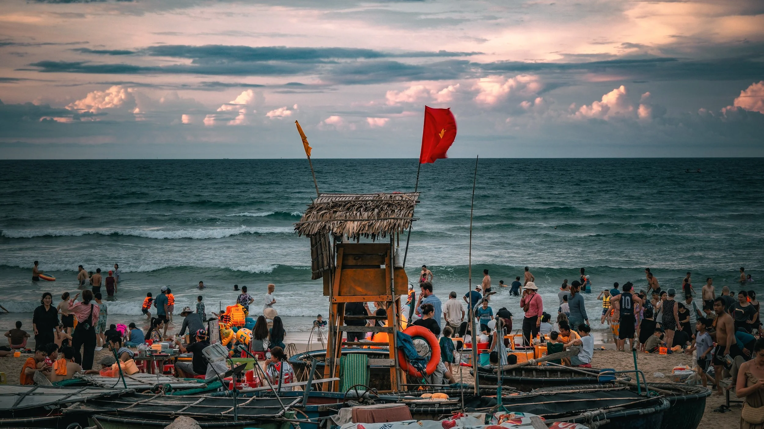 Crowded beach scene with people swimming and relaxing, colorful flags, a small overwater hut, and boats near the shore during sunset.