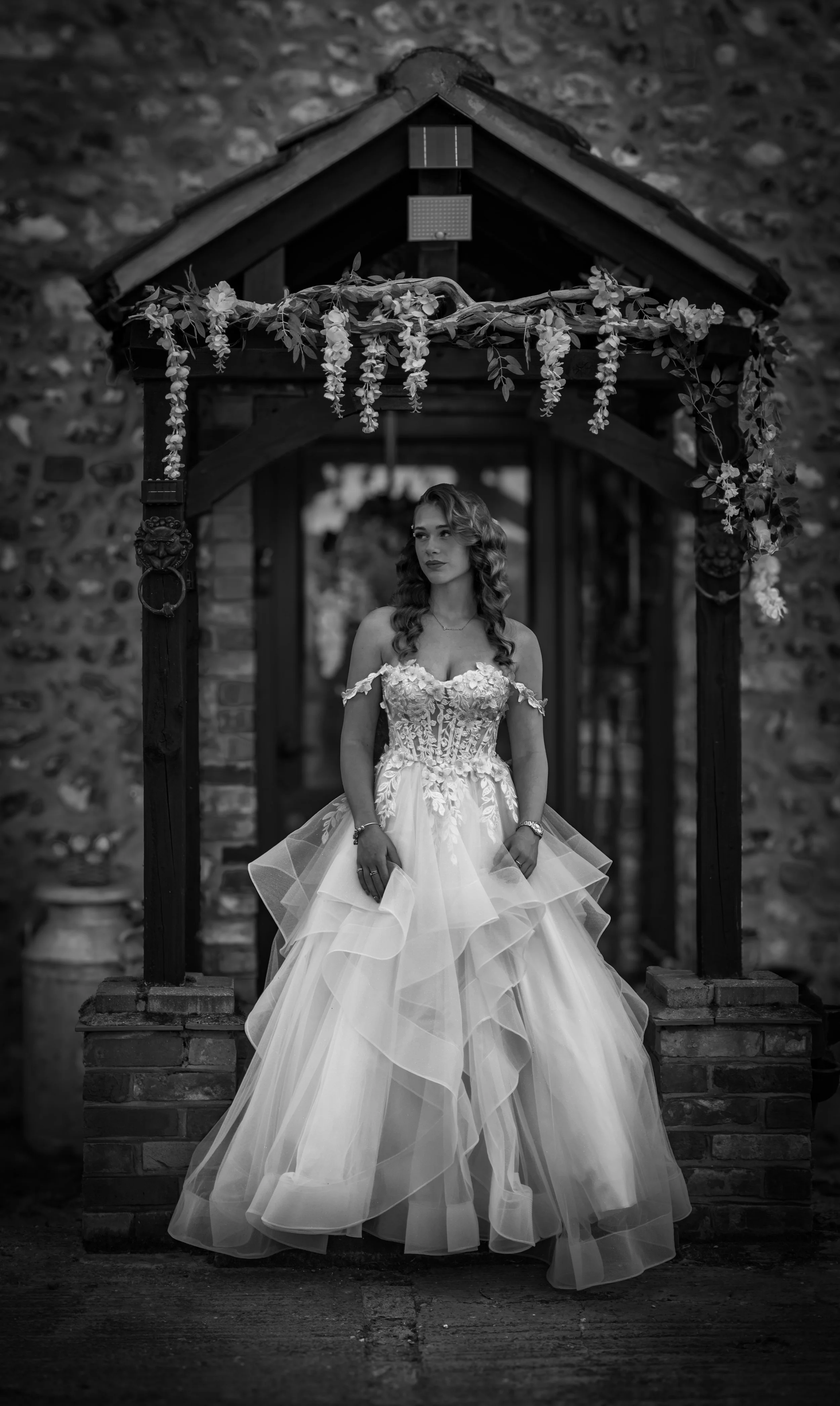 A woman in a wedding dress standing beneath a decorated archway with flowers, in front of a stone wall.