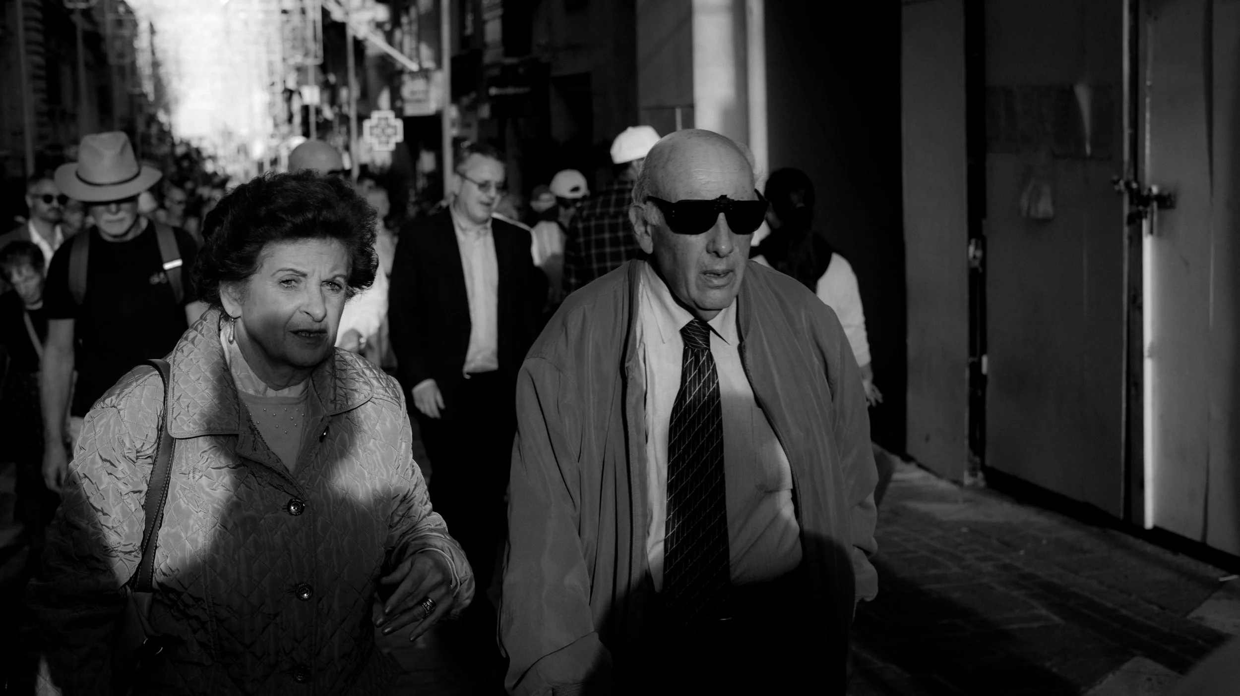 People walking on a busy city street in black and white, including an older woman with short dark hair and a light jacket, and a bald man with sunglasses, a striped tie, and a light-colored jacket.