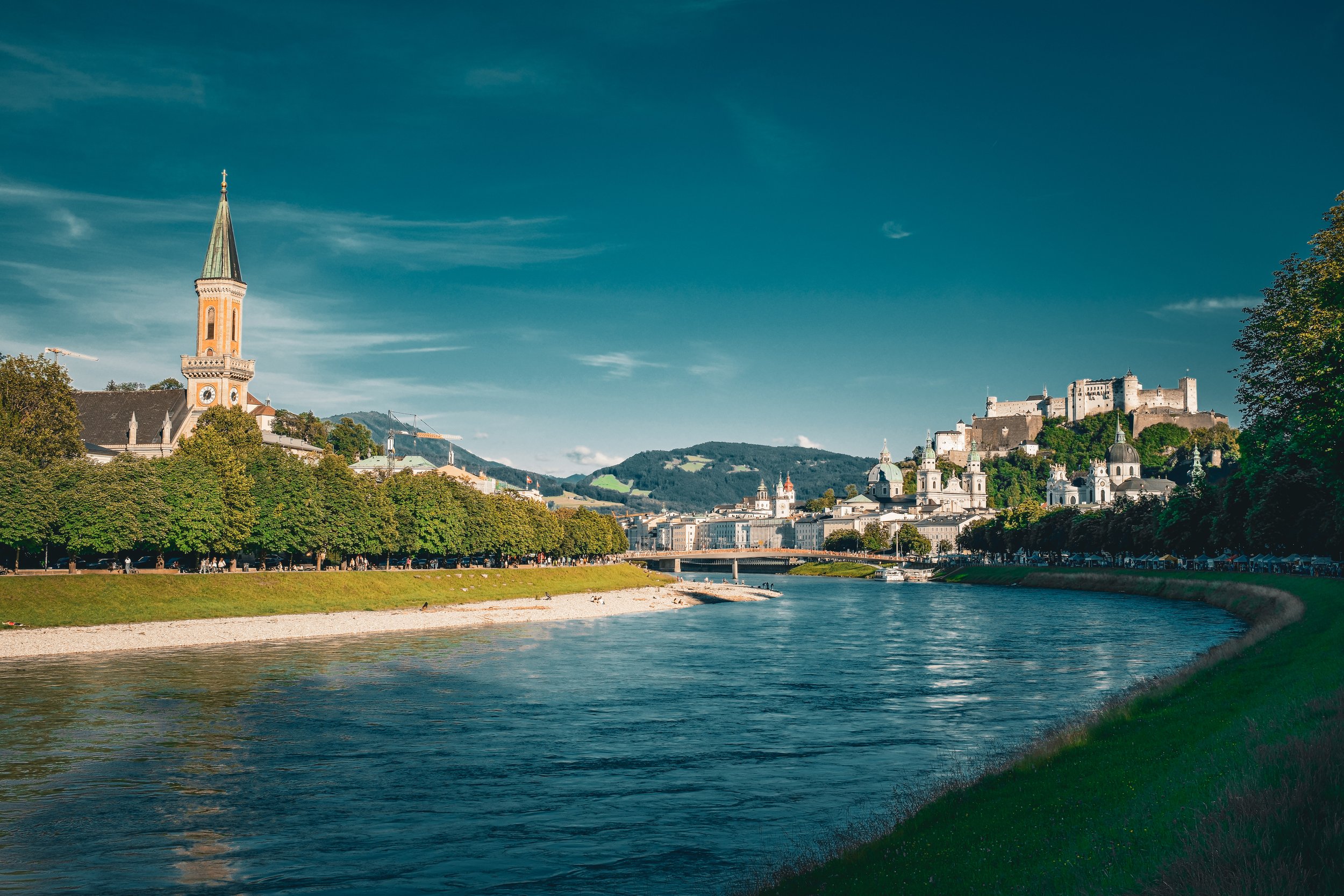 A scenic view of a river flowing through a city with historic buildings and a castle on a hill in the background under a clear blue sky.