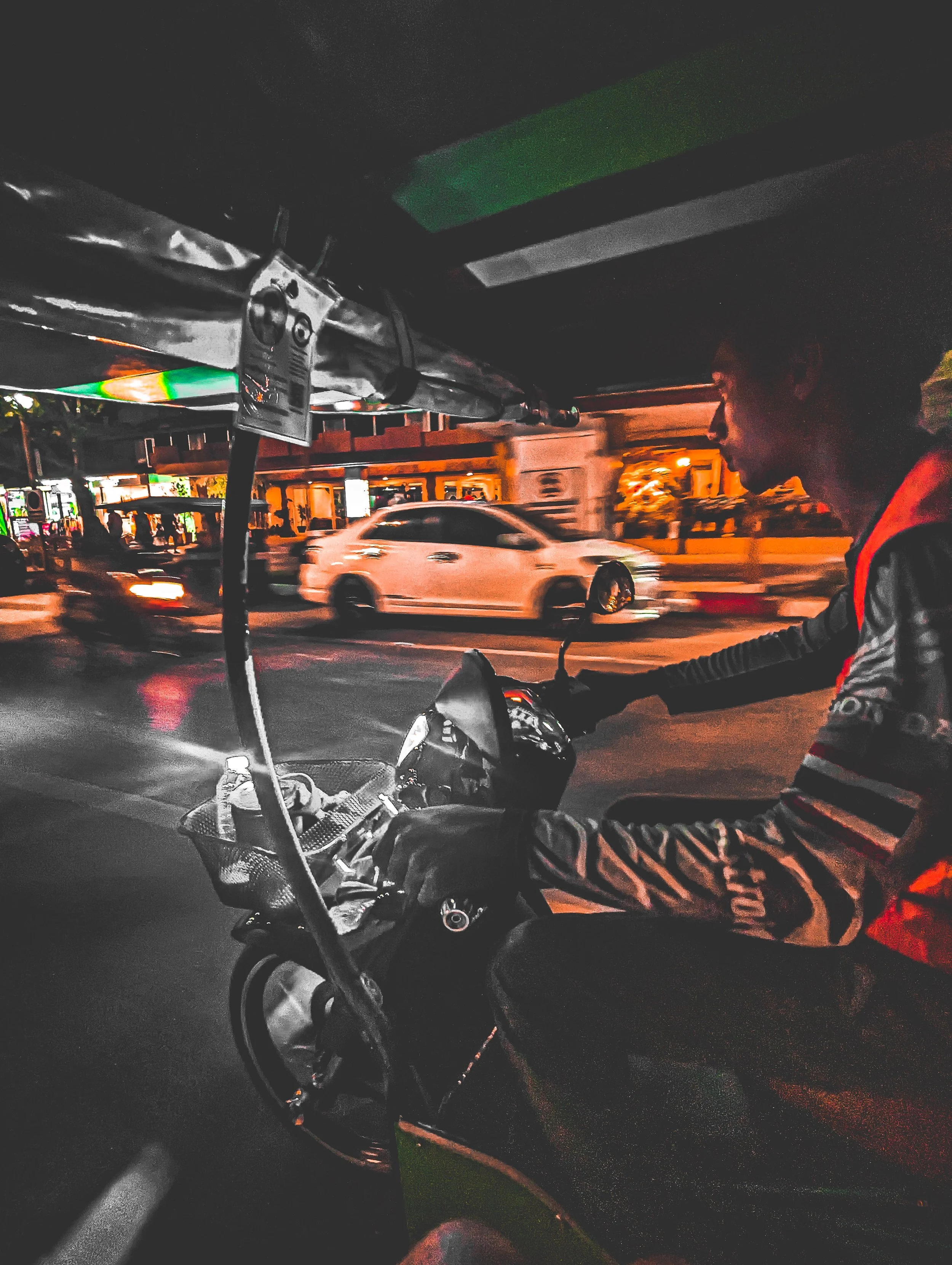 A person riding a motorcycle at night on a busy street. The person is wearing a jacket with reflective stripes and a red vest. Cars are visible on the road, and the street is illuminated by streetlights.