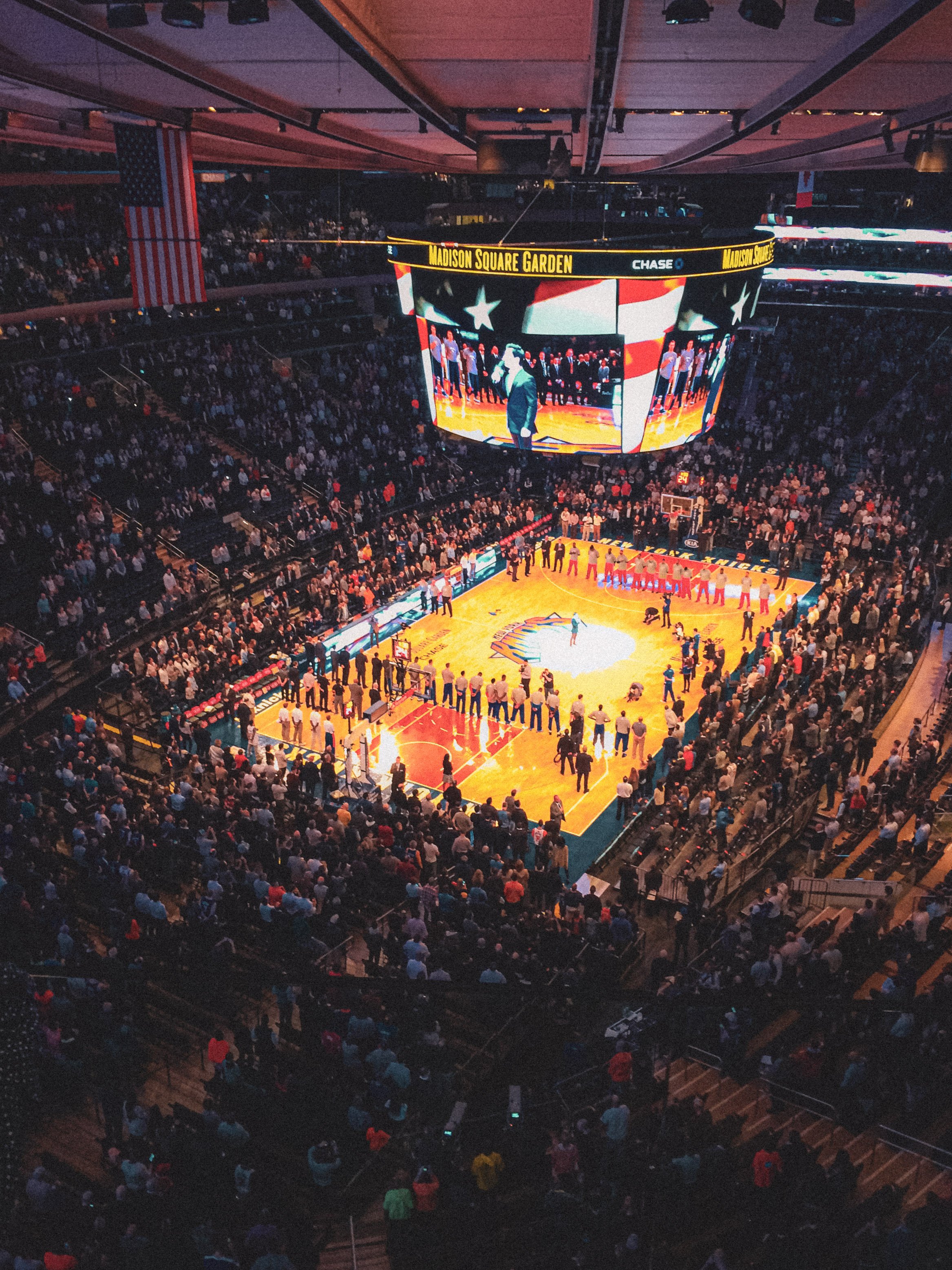 Crowd inside Madison Square Garden during a basketball game, with players on the court, and an electronic scoreboard displaying a speech or presentation.