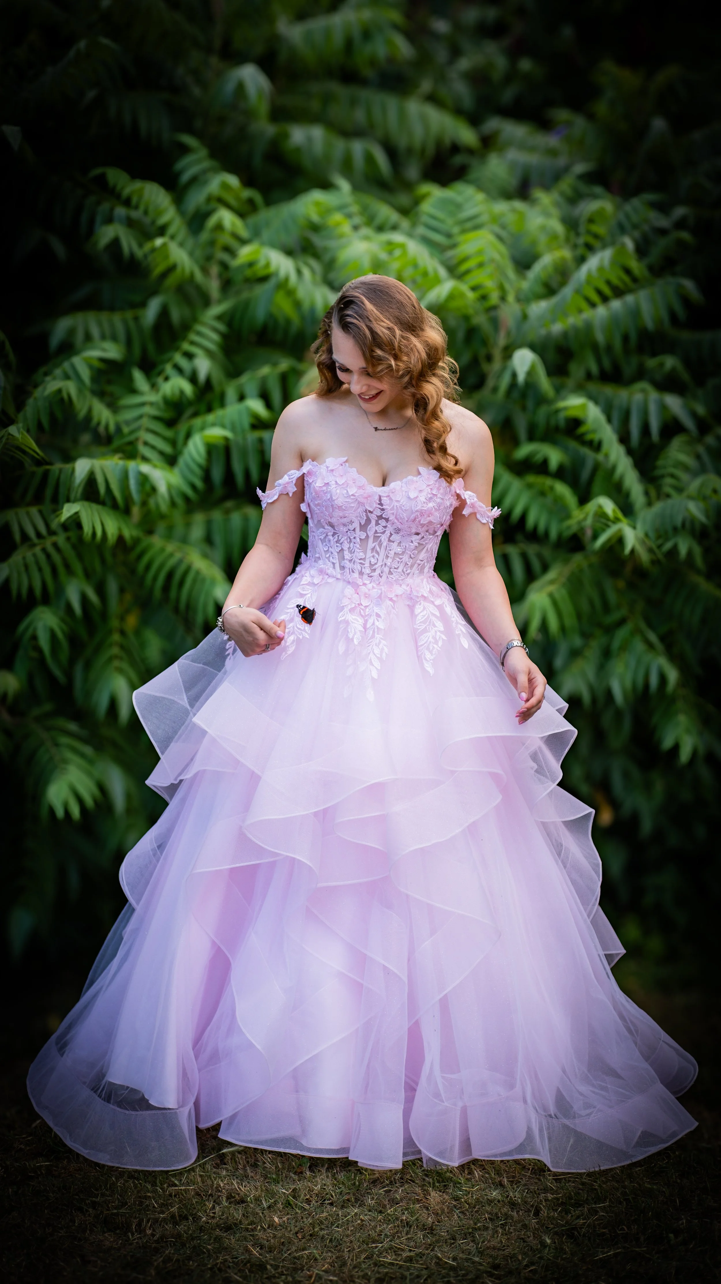 A woman in a pink strapless ball gown with lace details and layers of tulle is standing outdoors in front of lush green tropical plants, smiling and holding a butterfly on her finger.