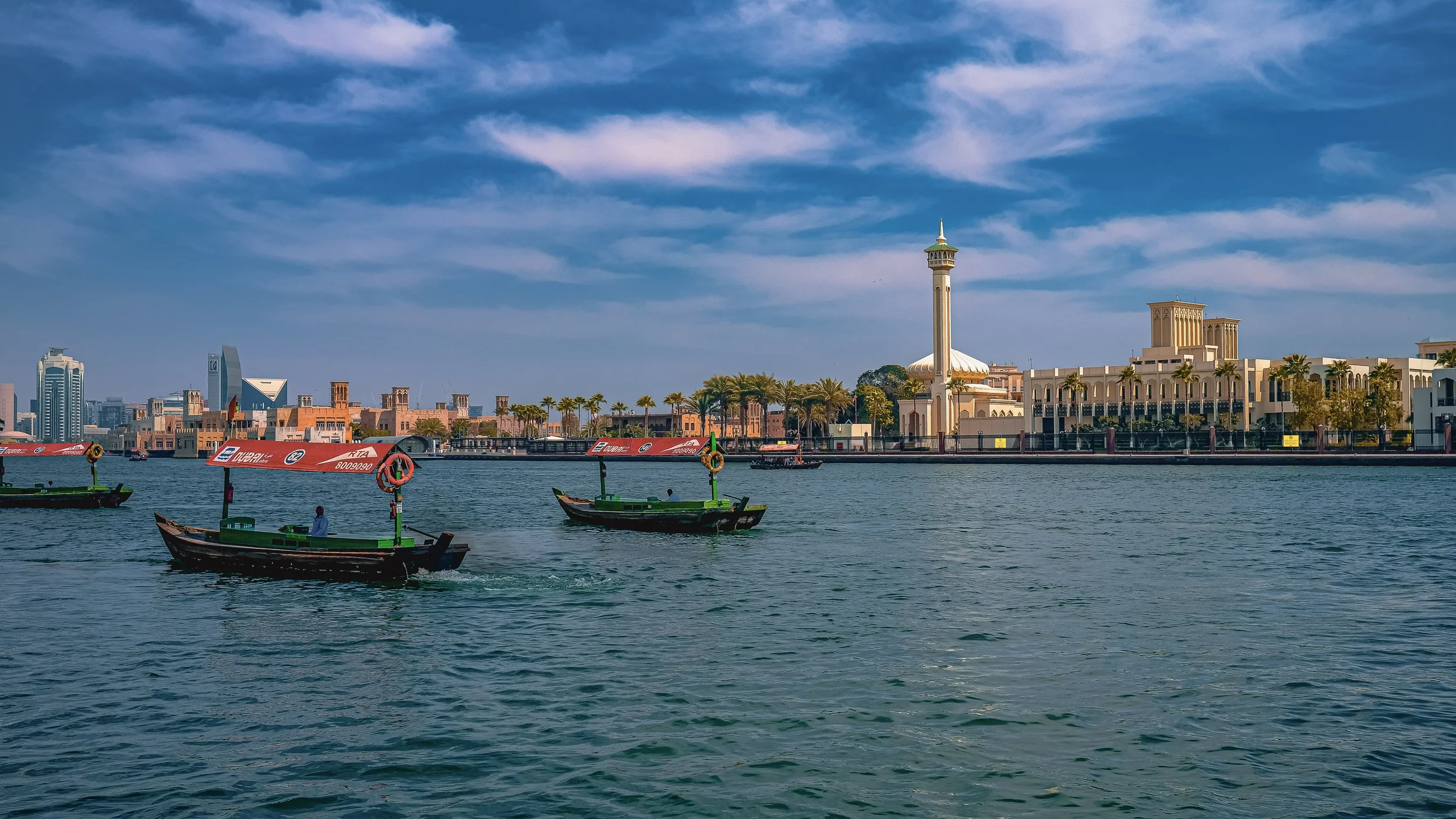 A view of a waterfront with boats on the water, including traditional wooden boats and modern boats, with a city skyline featuring tall buildings and a distinctive tower with a pointed top in the background. The sky is blue with some clouds.