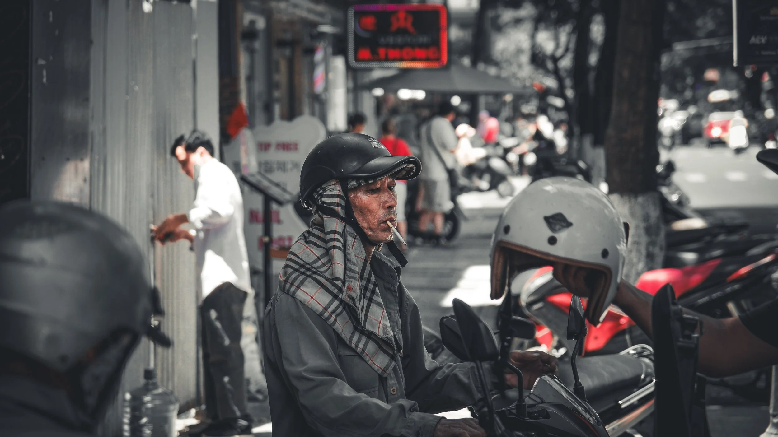An elderly man wearing a motorcycle helmet and a plaid scarf, sitting on a parked scooter at a busy street sidewalk, with blurred people and vehicles in the background.