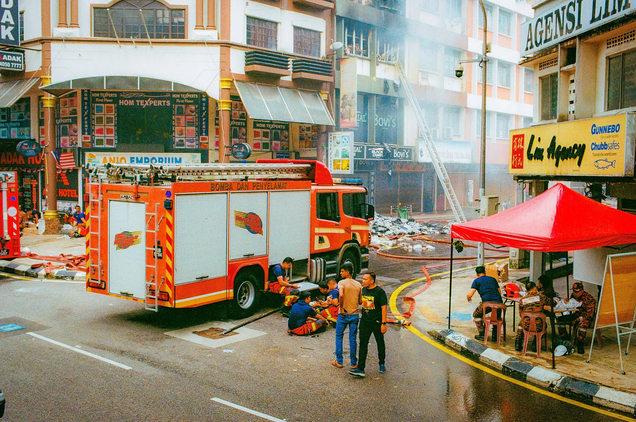 Emergency responders are attending to a fire incident on a busy urban street, with a fire truck and firefighters present, and smoke visible in the background.