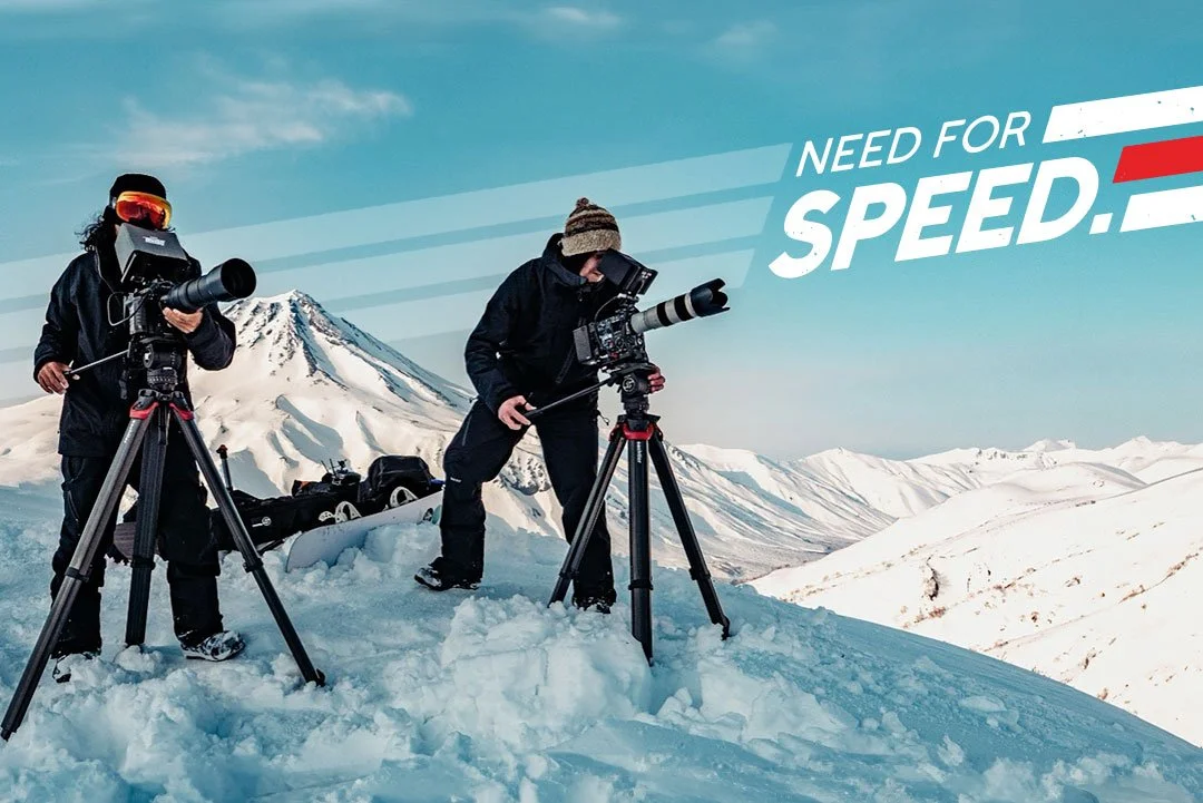 Two photographers in winter gear using cameras mounted on tripods on snowy mountain landscape with snow-capped peaks and a volcano in the background, with text "Need for Speed" in the sky.