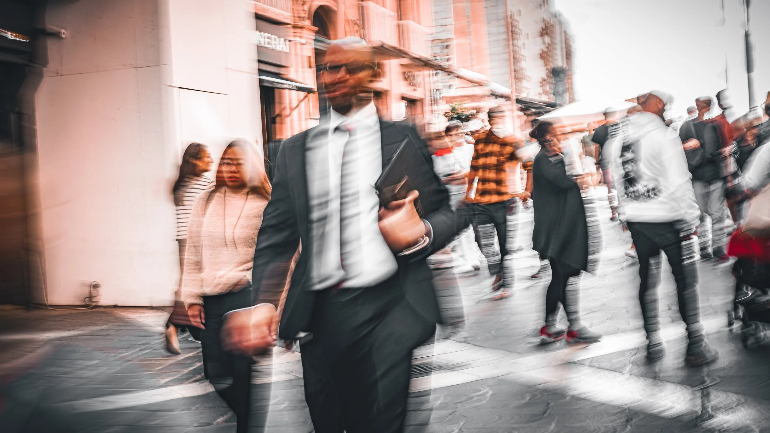Blurred image of people walking on a city street, with some gathered in a line, and buildings in the background.