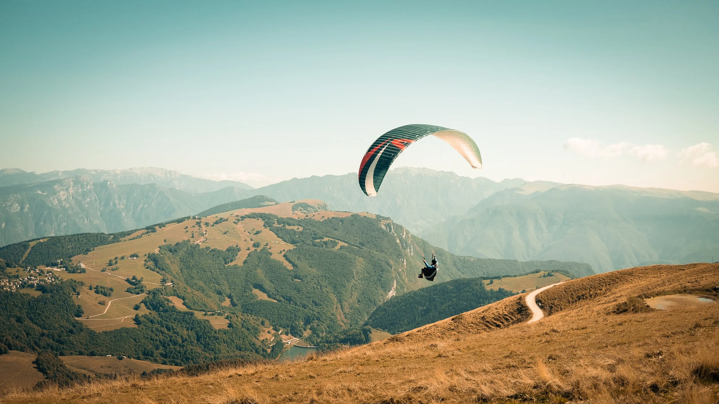 Person paragliding over a mountain landscape with green hills, winding paths, and distant mountains.