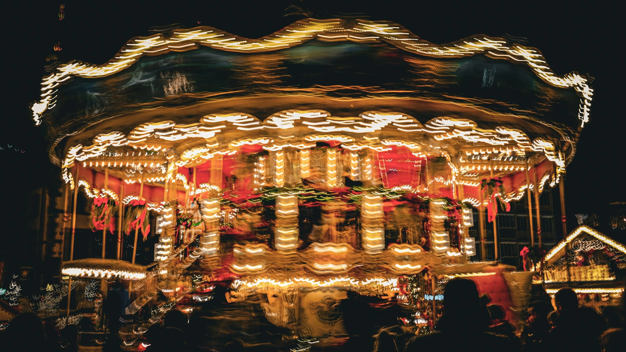 Blurred image of a merry-go-round with bright lights, red decorations, and children riding it at night.