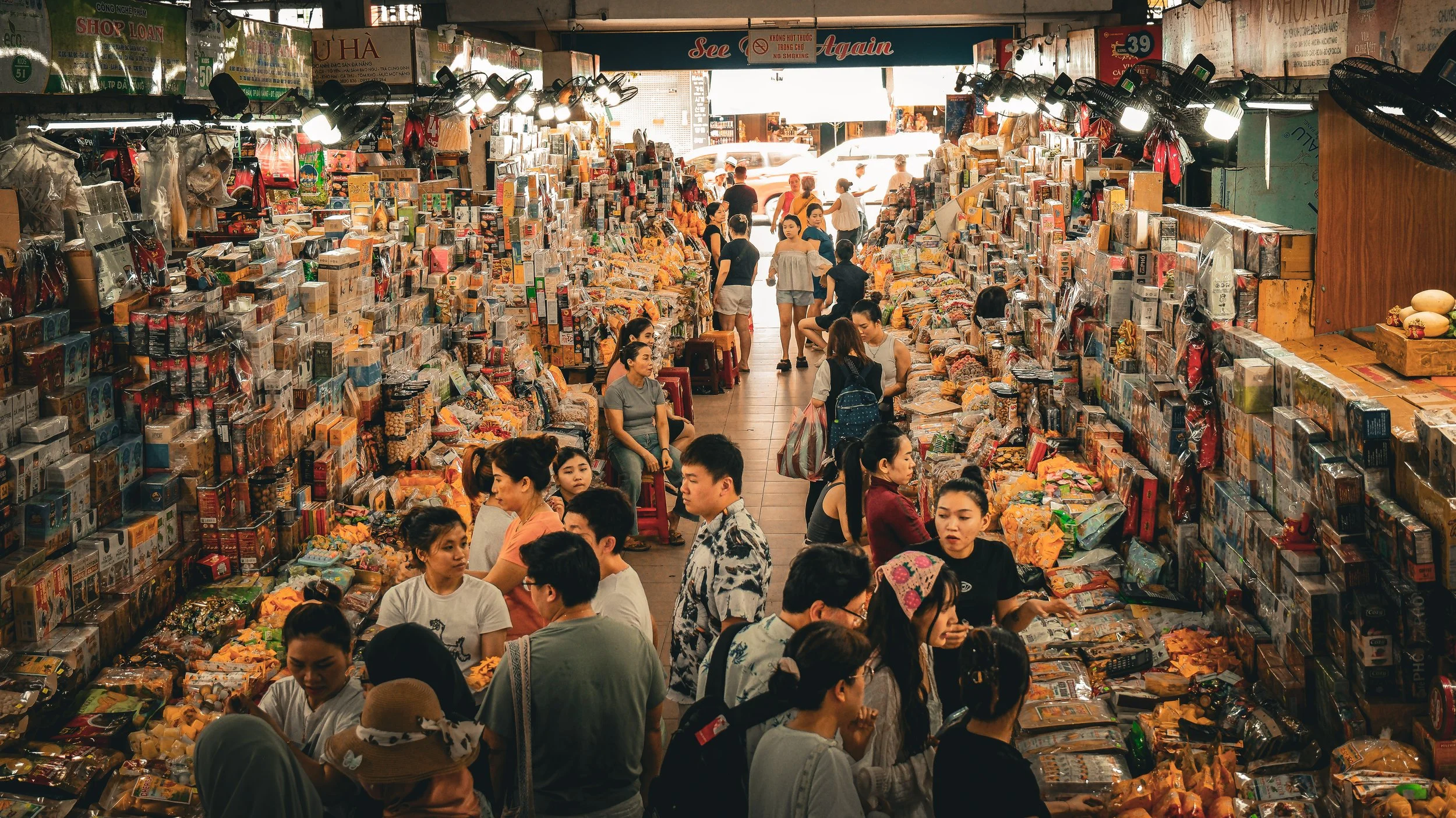 A busy indoor market with shelves filled with goods, many shoppers browsing and sitting, with sunlight coming through the open entrance in the background.