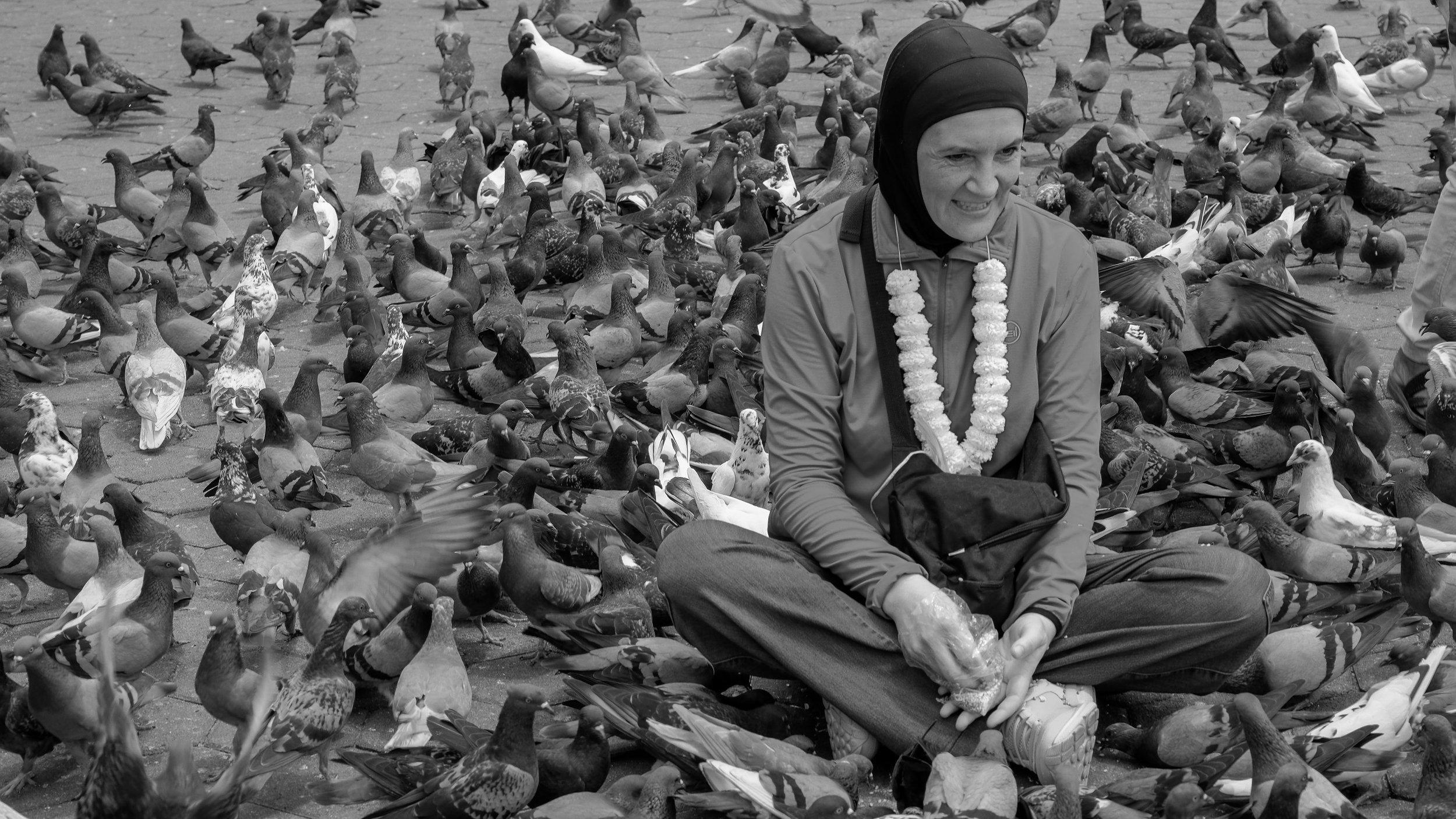 A woman sitting on a paved surface surrounded by numerous pigeons. She is wearing a head covering and a scarf decorated with pom-poms, and appears to be smiling or grimacing. The pigeons are both on the ground and flying.