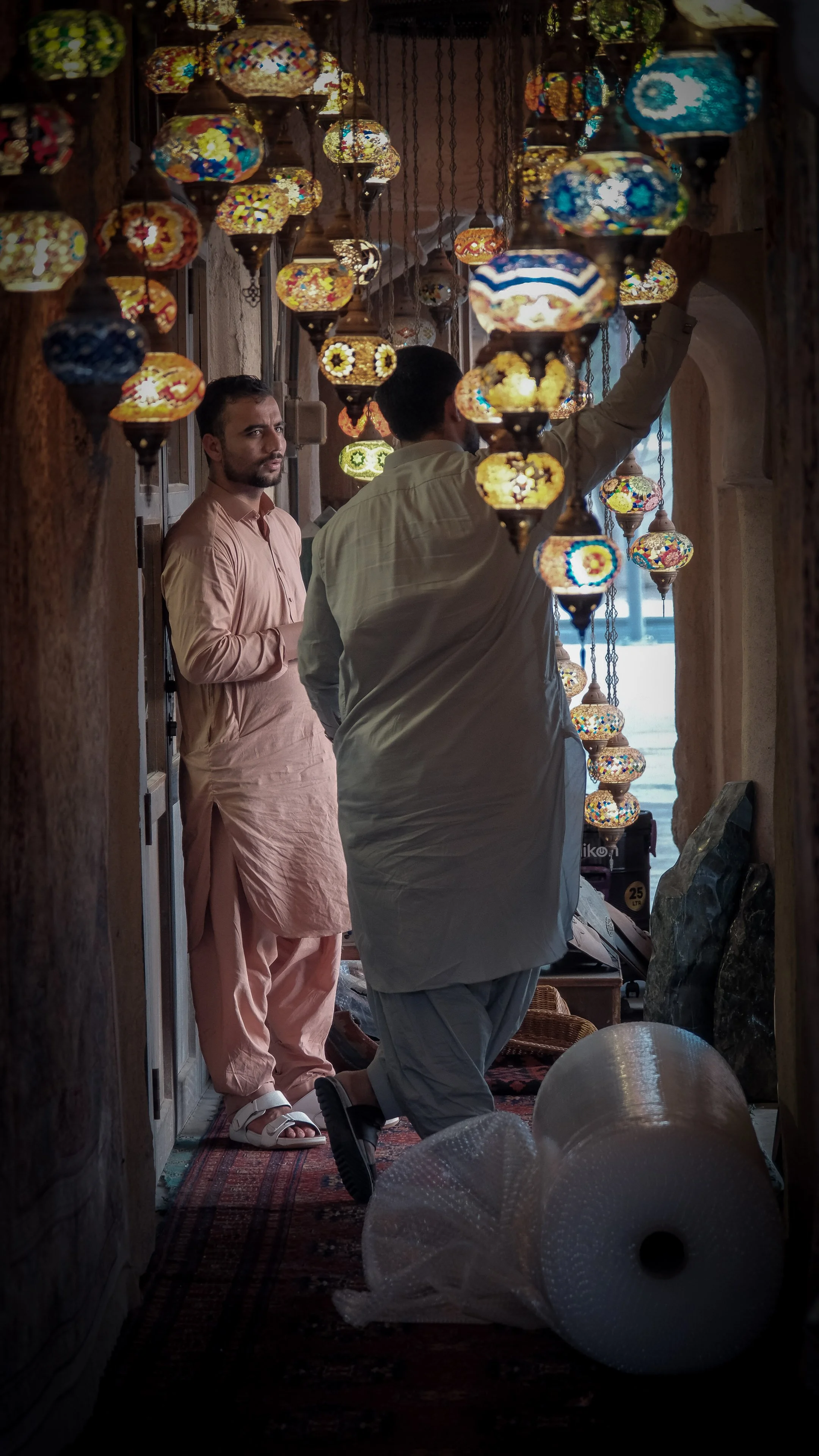 Two men in traditional clothing, one adjusting colorful mosaic lamps hanging from the ceiling, inside a warmly lit room with ornate decor.