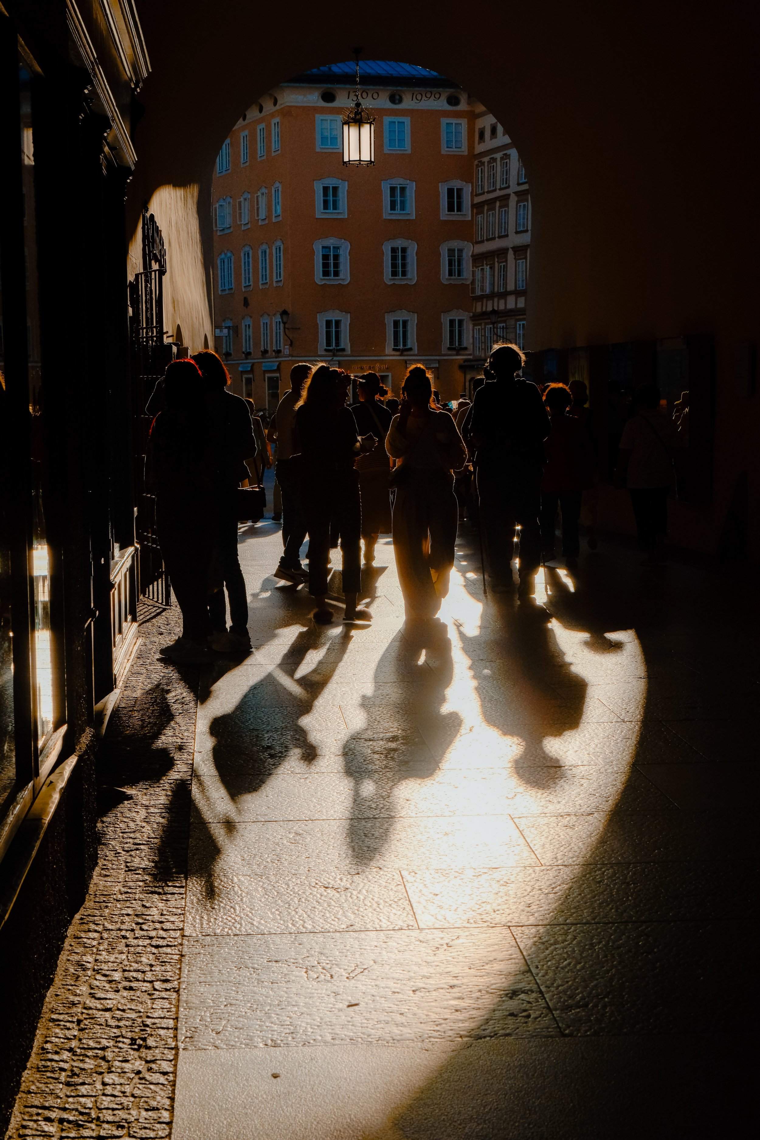 People walking through a tunnel with sunlight casting shadows on the ground, and a building with multiple windows visible in the background.
