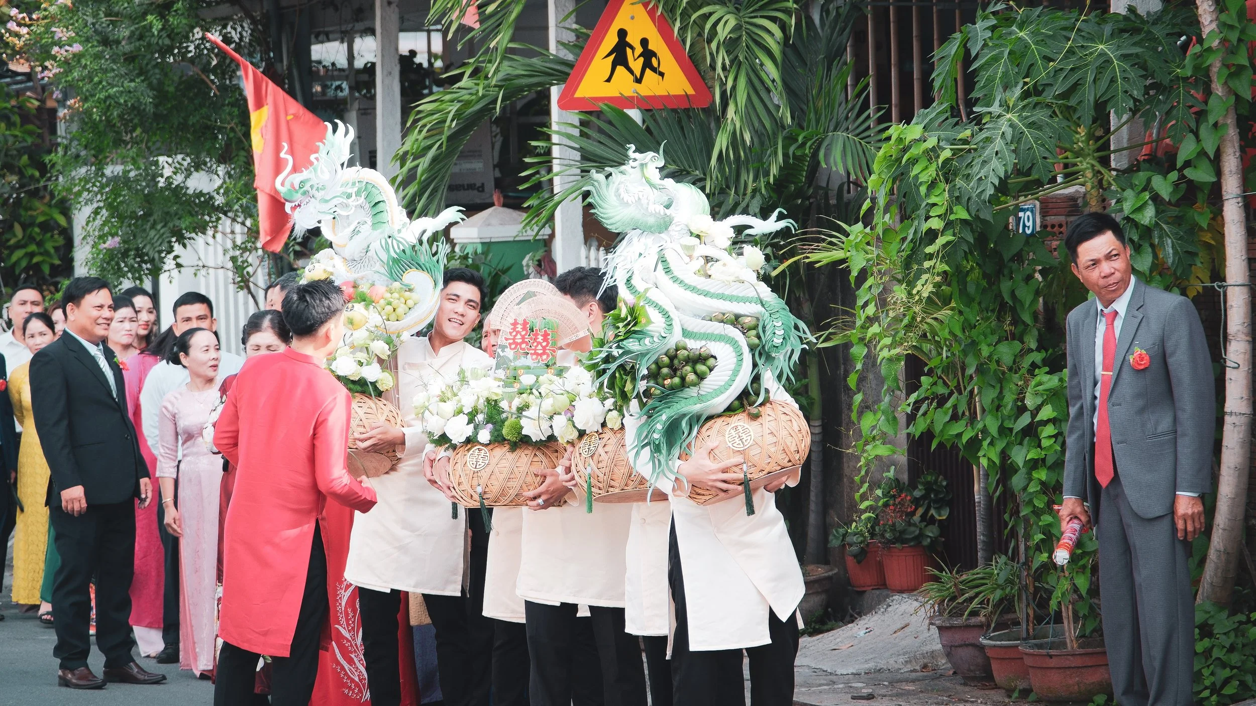 A traditional Vietnamese wedding scene with a group of people, including men and women in formal and traditional attire, on a street. Several large decorative dragon sculptures with flowers are carried by people in white outfits. A man in a gray suit