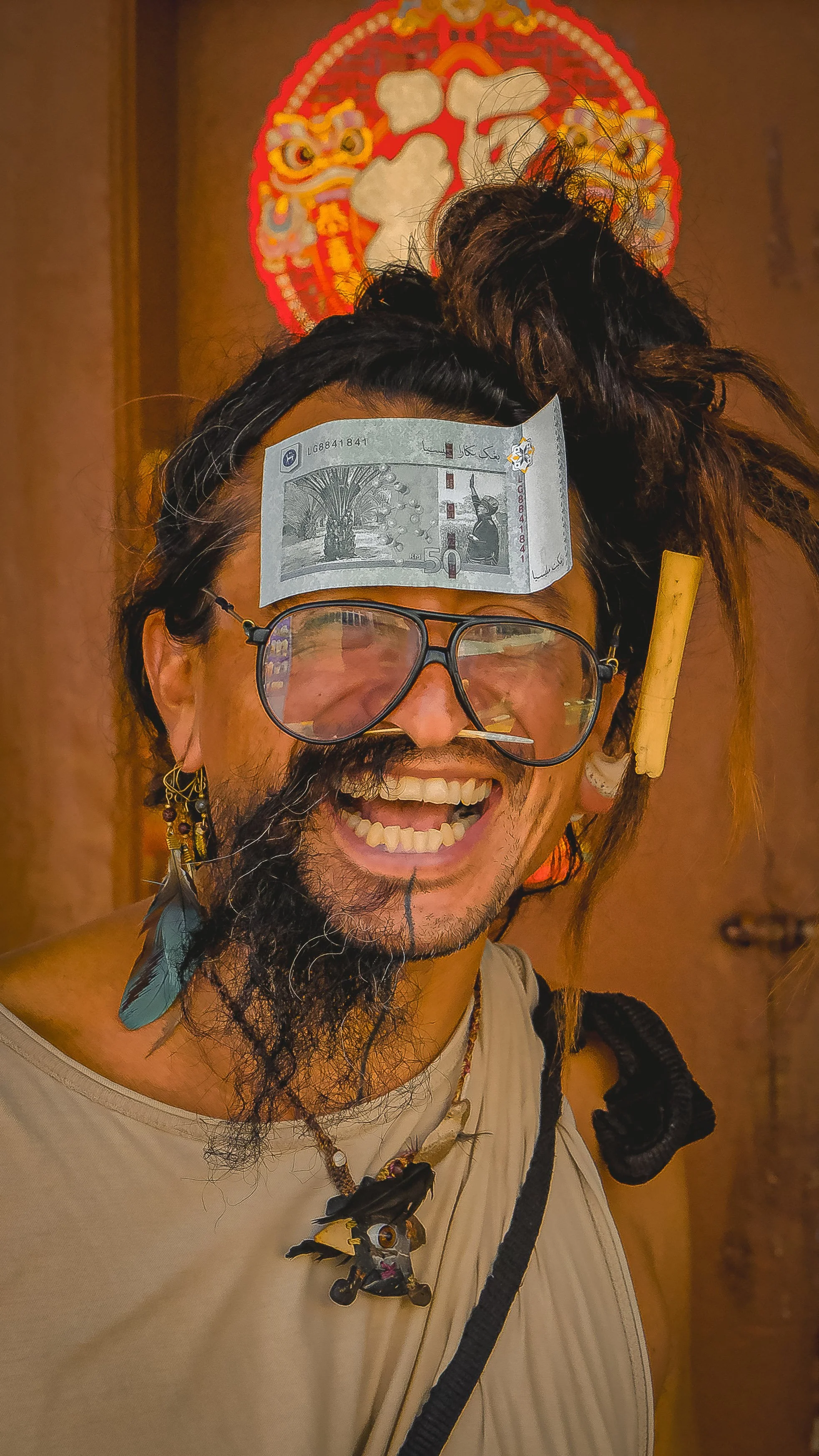 A person with dreadlocks and glasses smiling, with a fifty ringgit banknote on his forehead, a yellow rolled paper in his hair, and a dragon head artwork in the background.