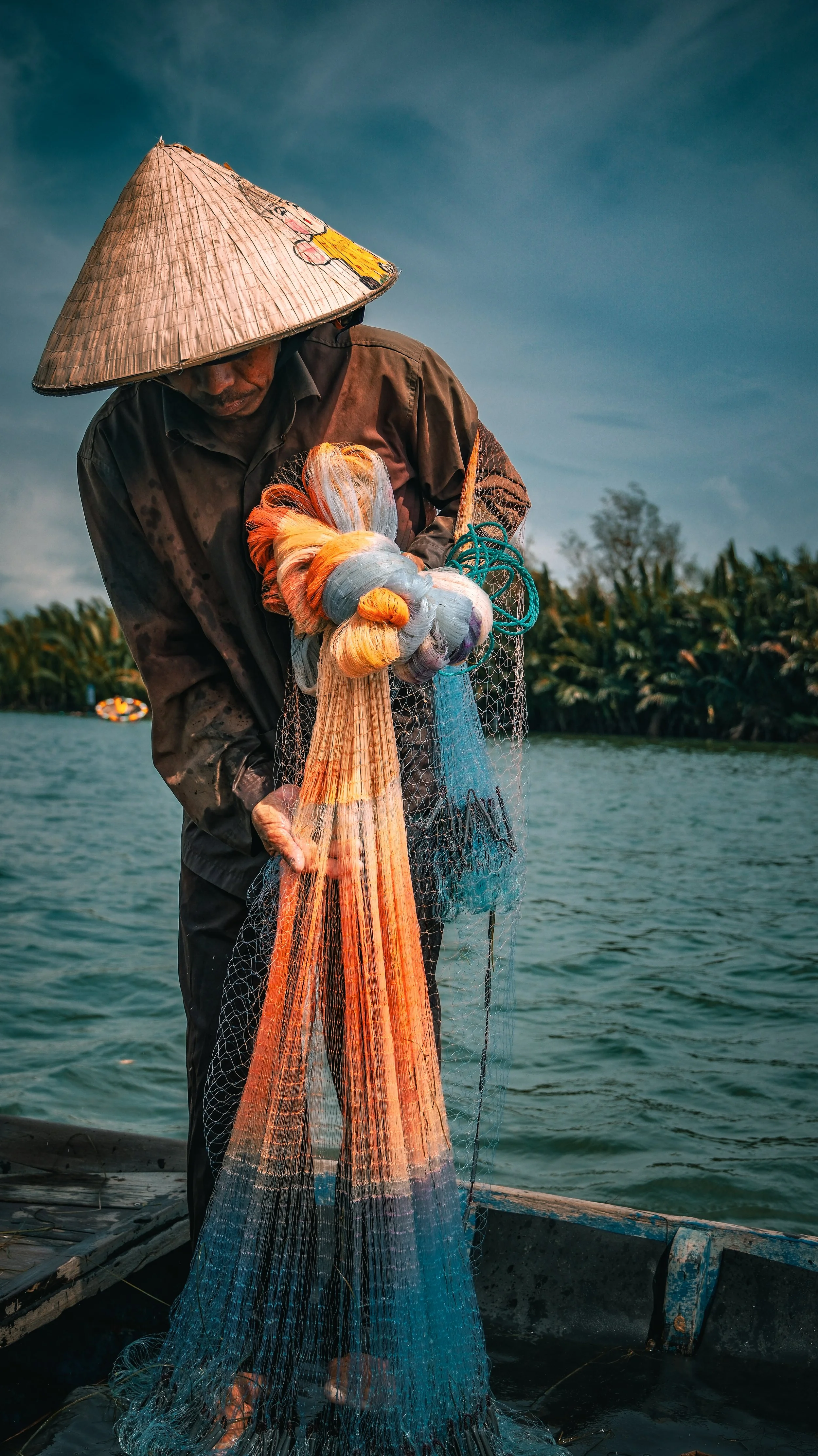 A fisherman wearing a traditional conical hat stands on a boat, holding a colorful fishing net over water with lush greenery in the background.
