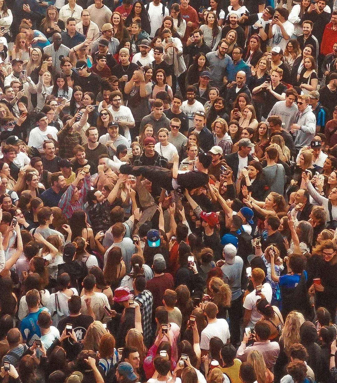 Large crowd of people gathered outdoors, many holding up smartphones, some taking pictures or recording. Some individuals are holding or raising objects, while others are clapping or watching attentively.