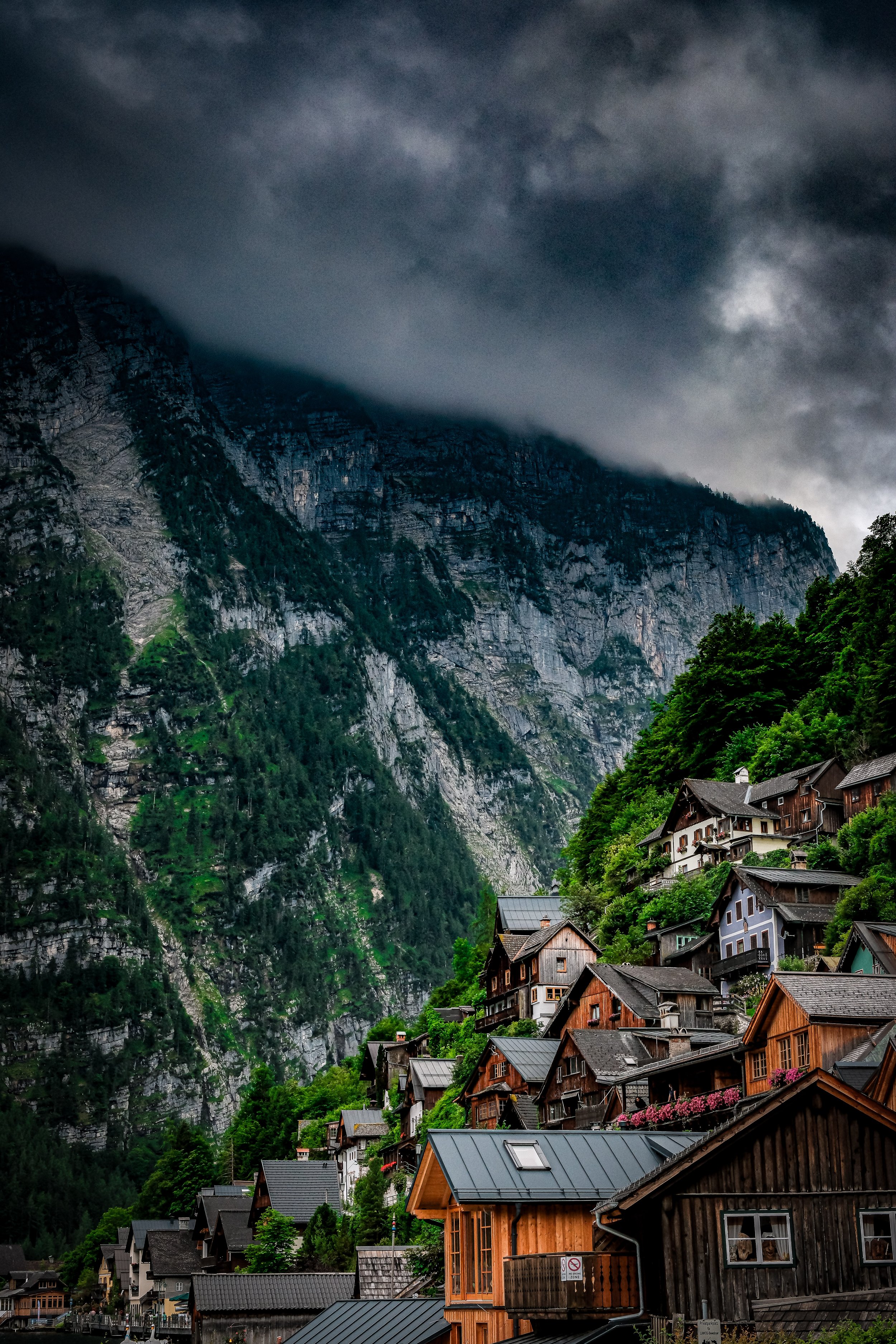 A mountain village with wooden houses on a steep hillside, lush green trees, and dark storm clouds overhead.