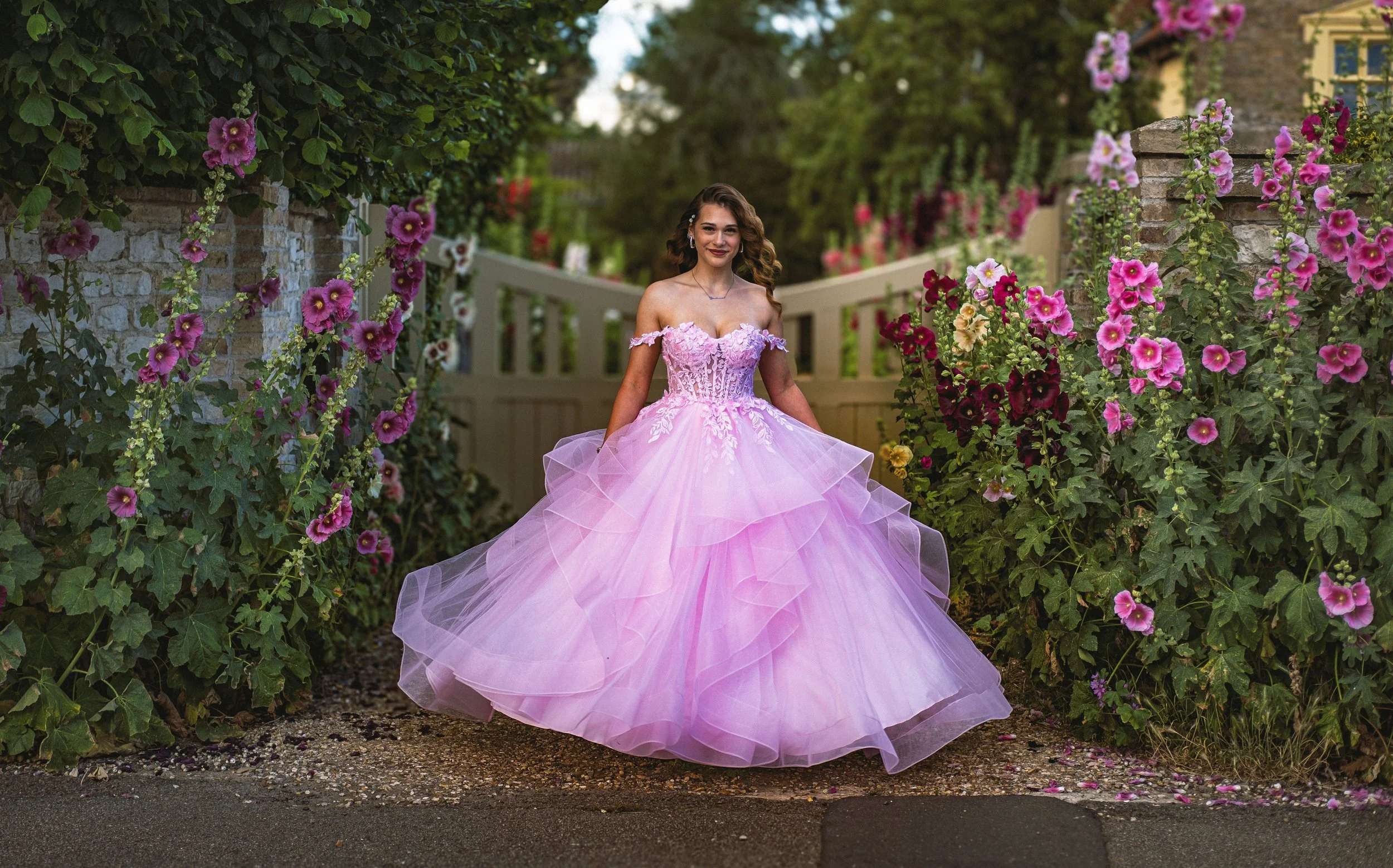 A young woman in a pink ball gown with floral embellishments is standing on a garden path surrounded by pink and purple flowers.