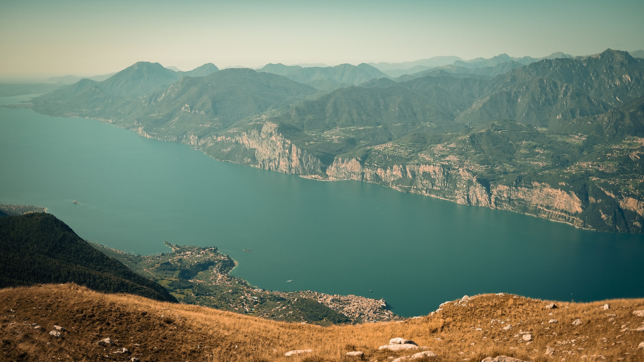 Aerial view of a large lake with mountainous terrain surrounding it, small settlements along the shoreline, and lush green forests in the foreground.