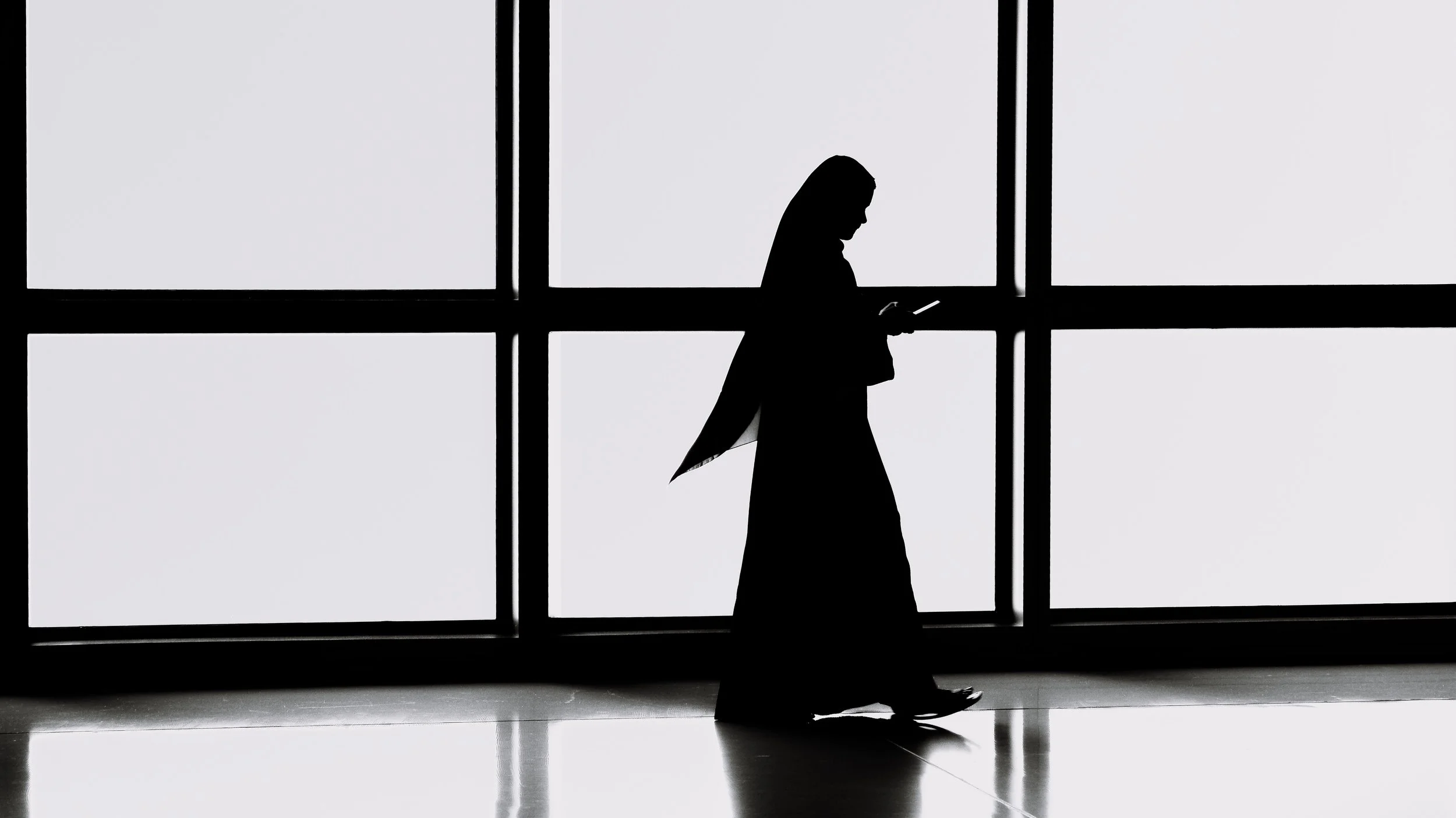 Silhouette of a woman walking while looking at her phone in front of large windows.