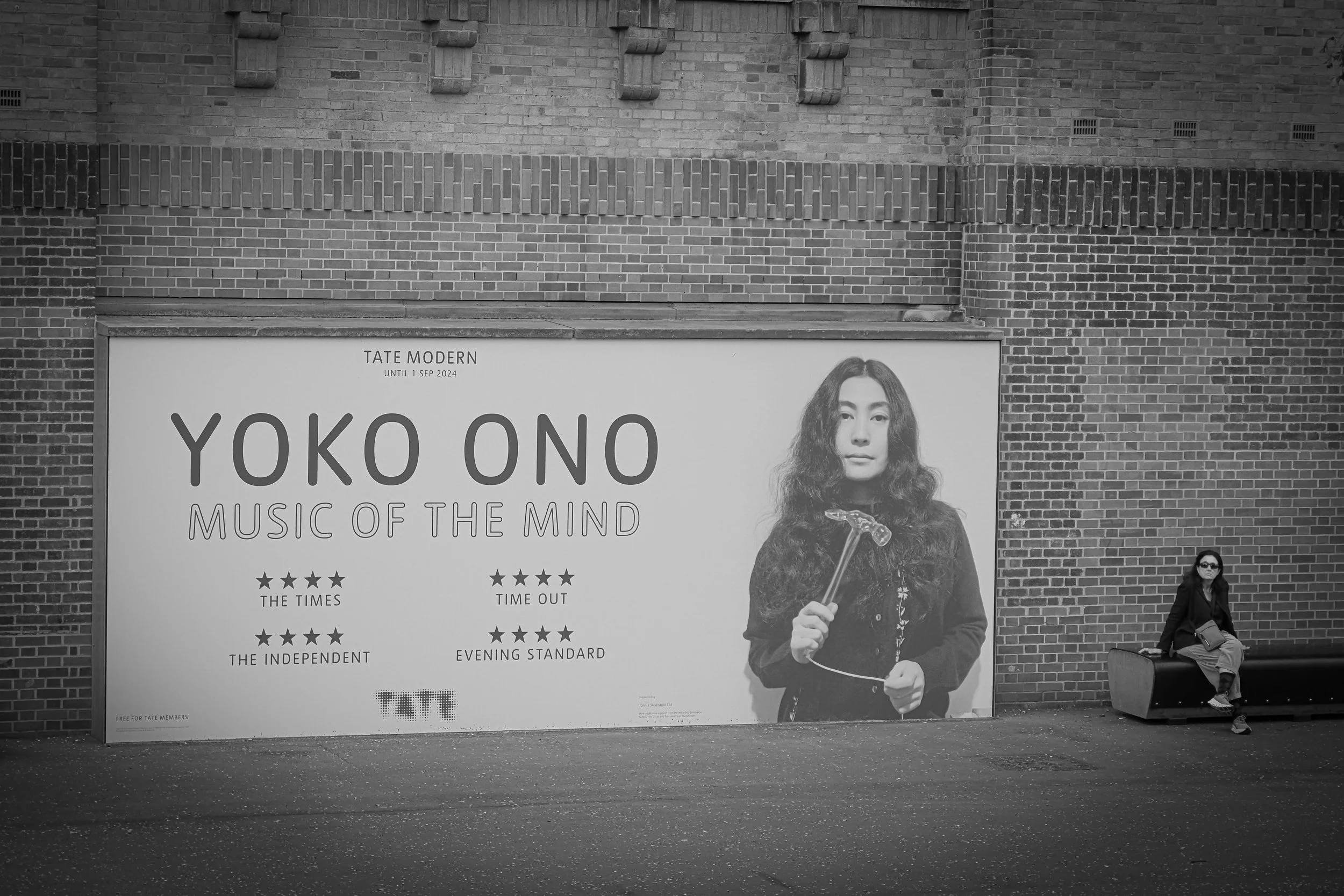 Black and white street scene with a large advertisement poster on a brick wall promoting Yoko Ono's music exhibit, featuring a portrait of Yoko Ono holding an object in her right hand. A woman in dark clothing and sunglasses is sitting on a bench bes