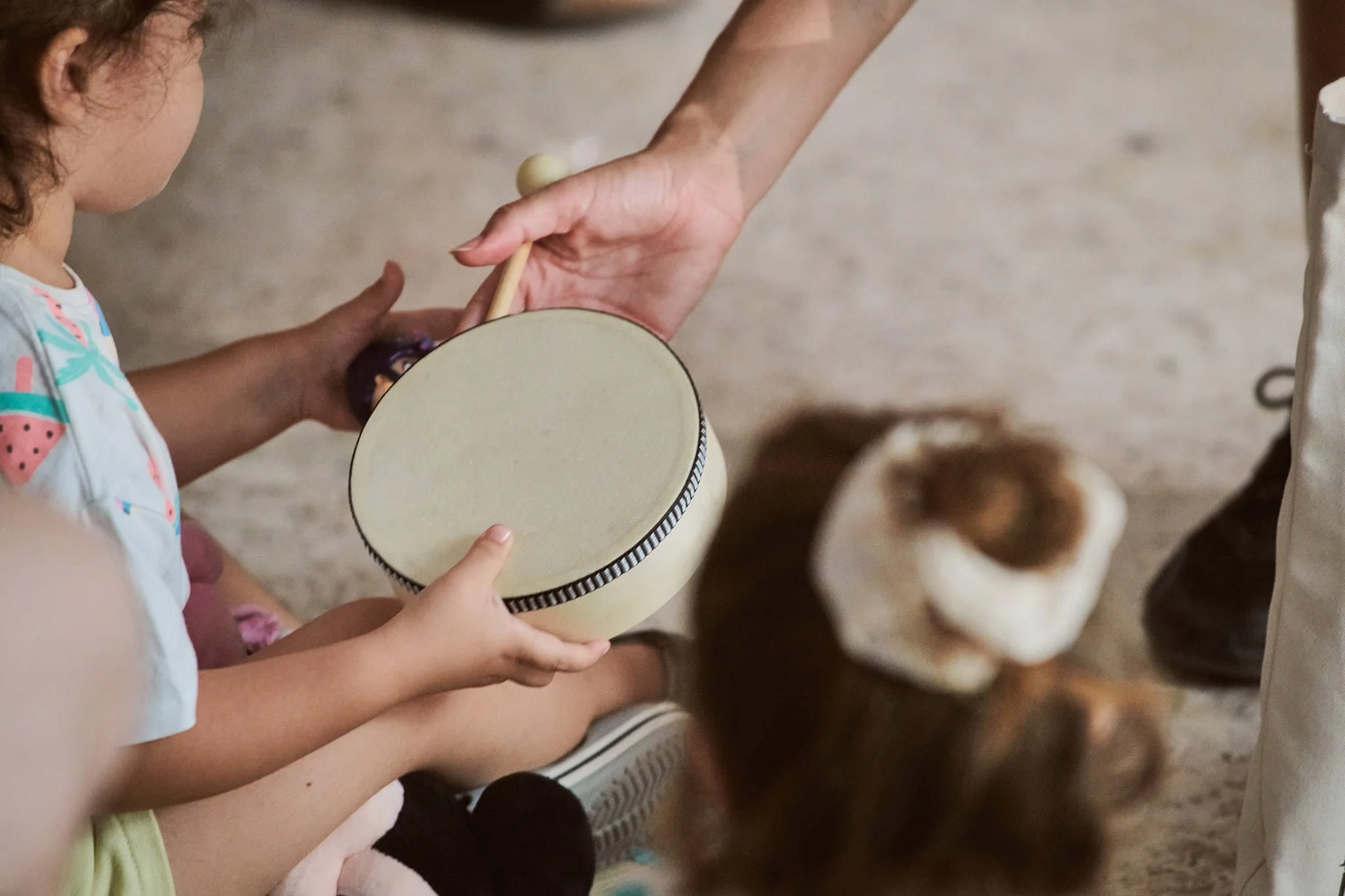 A child holding a drum with both hands in front of another child, who is partially visible. There are several dogs in the background.