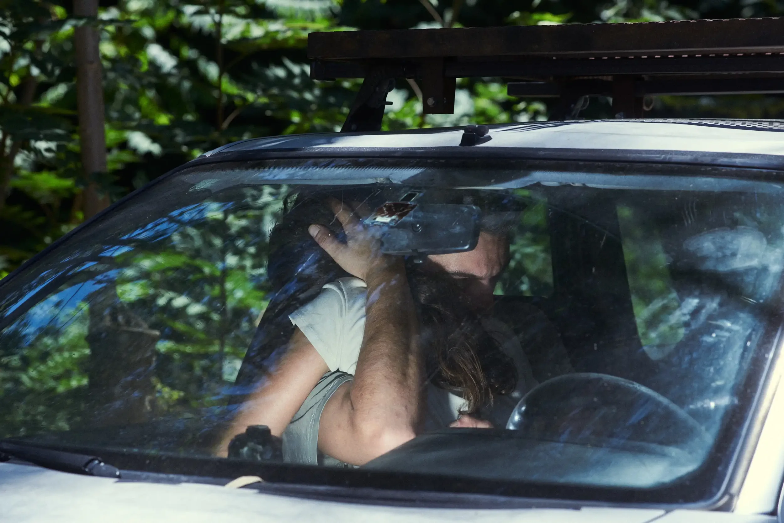A person sitting inside a vehicle, holding a cellphone to their ear, with reflective glass showing trees outside.