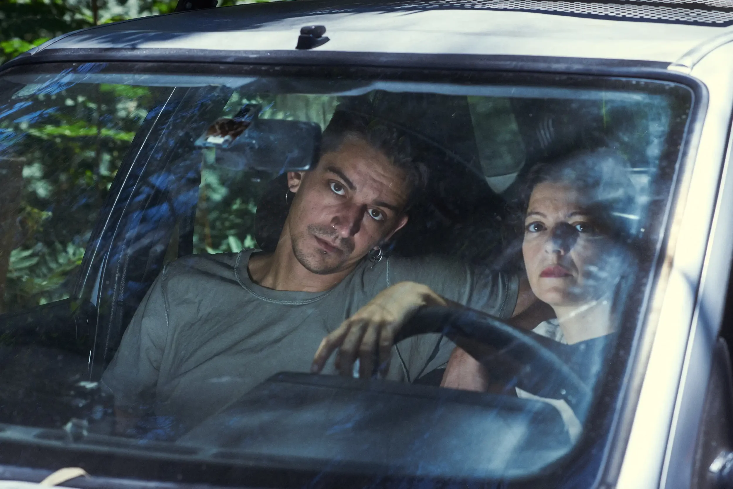 A man and woman sitting inside a vehicle, looking through the windshield with serious expressions.