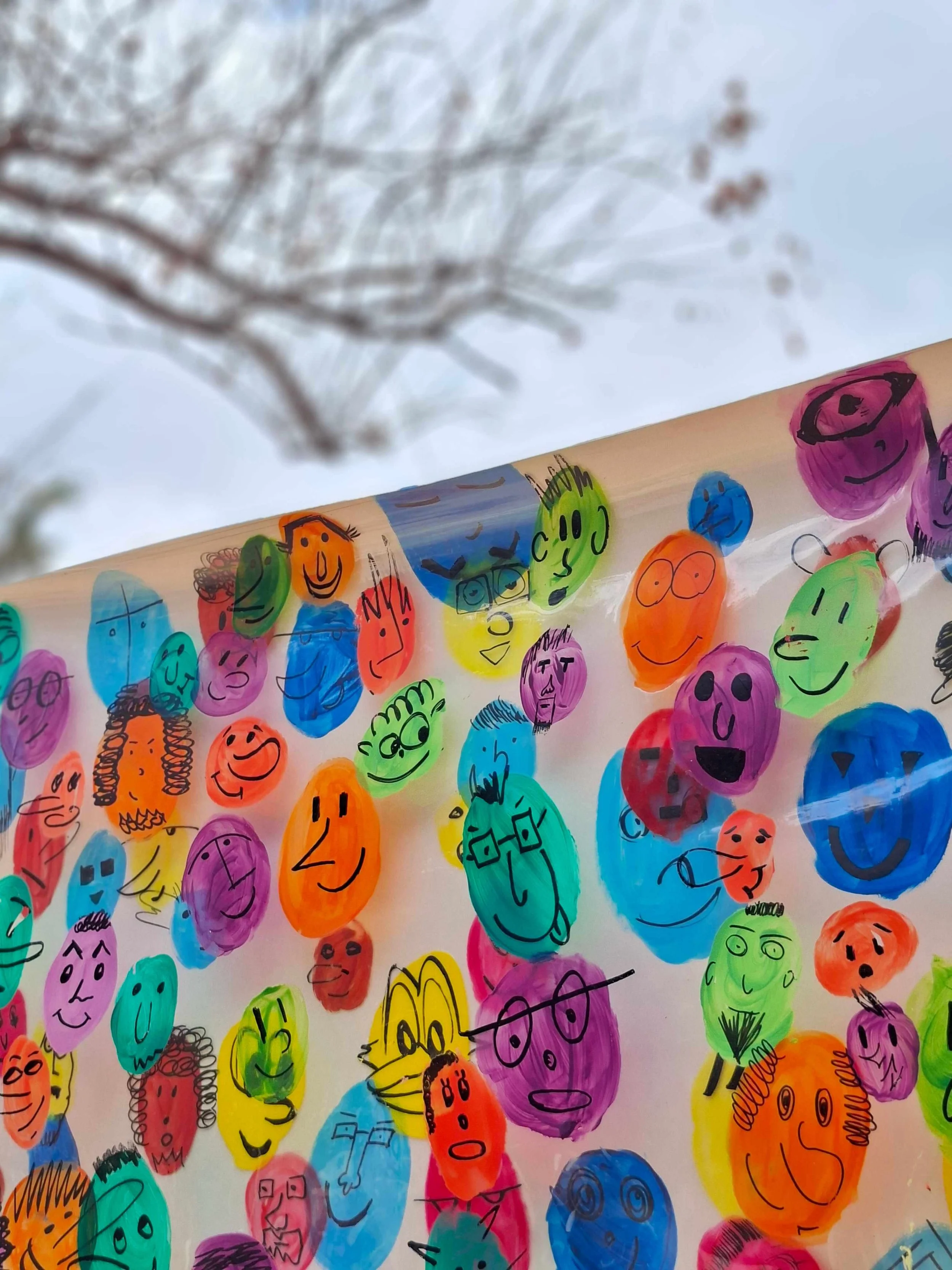 Close-up of a colorful paper with many hand-drawn smiley faces in various colors and sizes, with a background of blurred tree branches and sky.
