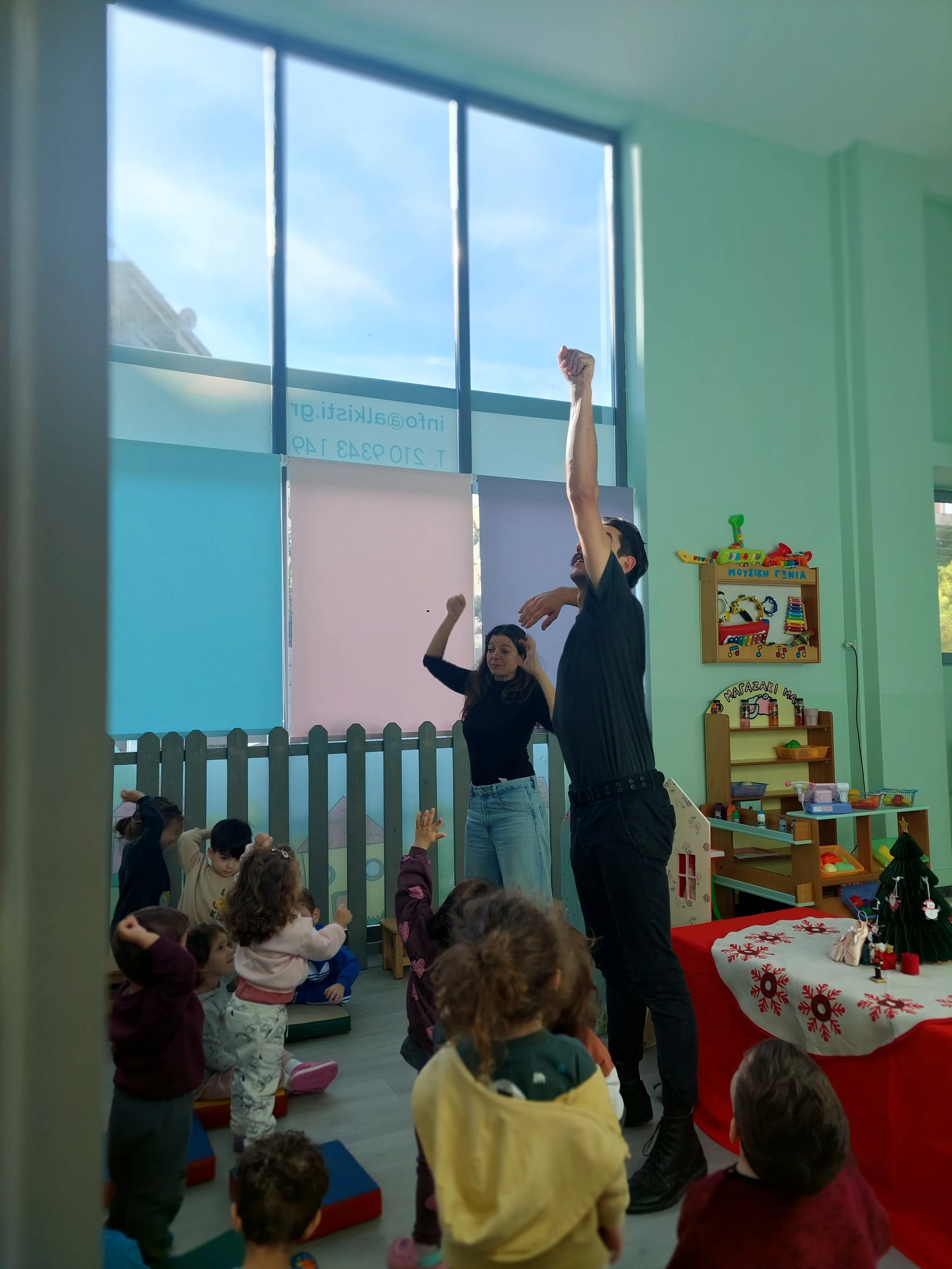 Children sitting and standing in a classroom, participating in a festive activity with two women. The room has large windows, toys, and holiday decorations.