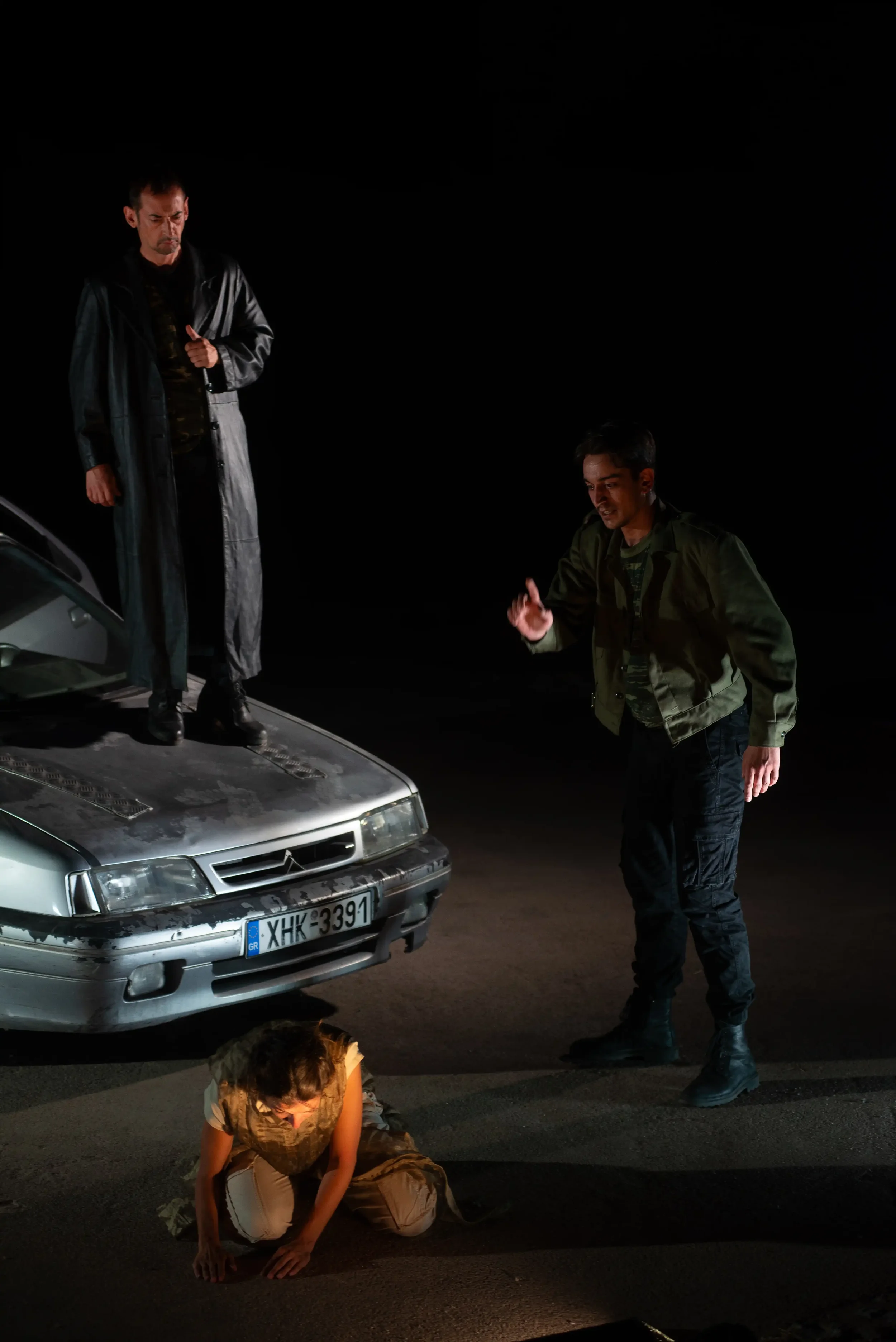 Three people are involved in a staged scene at night, with two men standing and one woman kneeling on the ground, next to a silver car with a Greek license plate. The scene is dark, with minimal lighting highlighting the individuals and car.