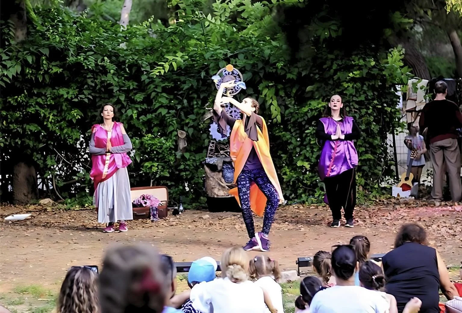 Performers on stage entertaining an audience in an outdoor setting, with lush green foliage in the background.