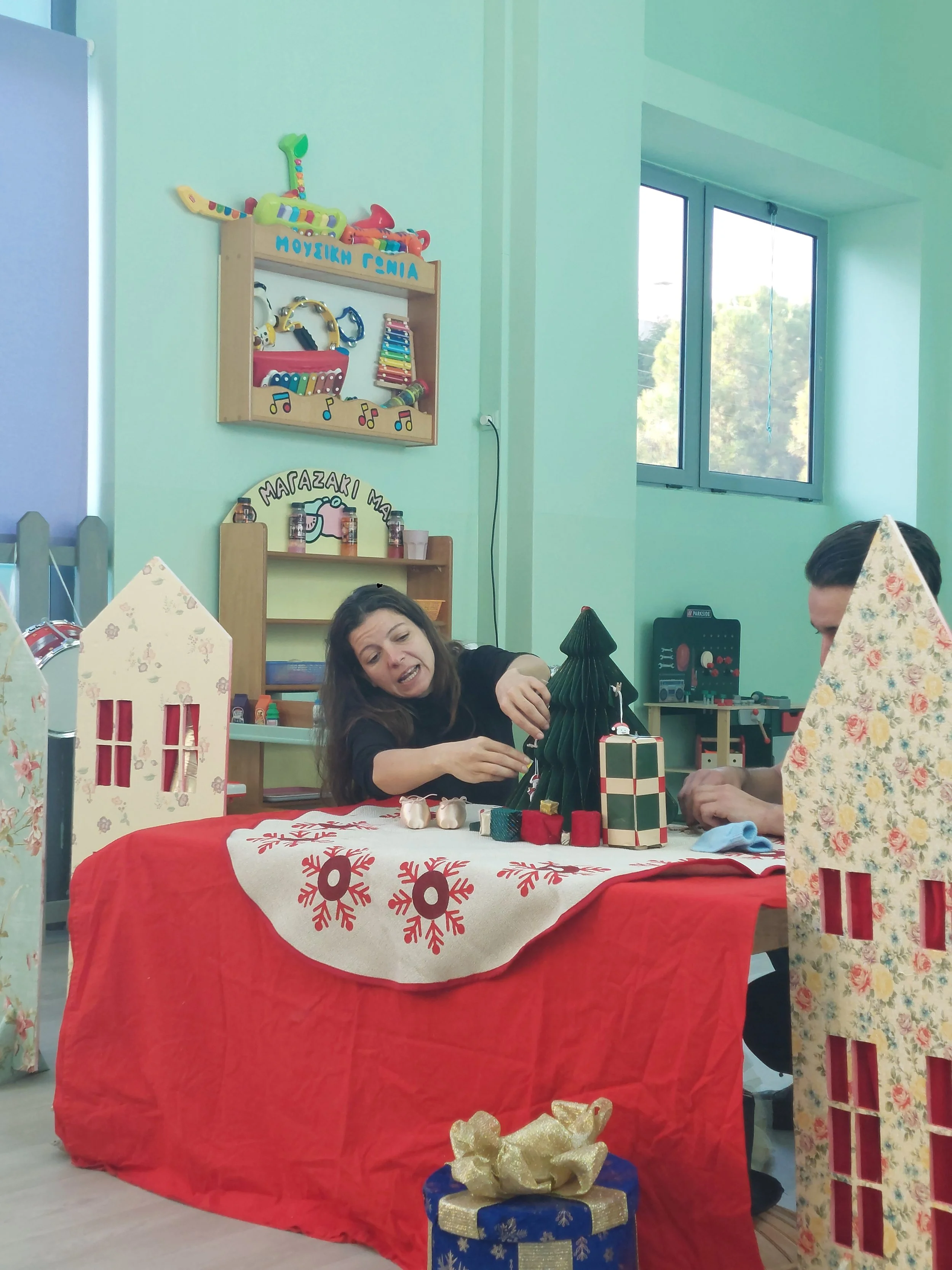 People decorating Christmas-themed items at a table with a red tablecloth and festive decorations, with holiday presents and window in the background.