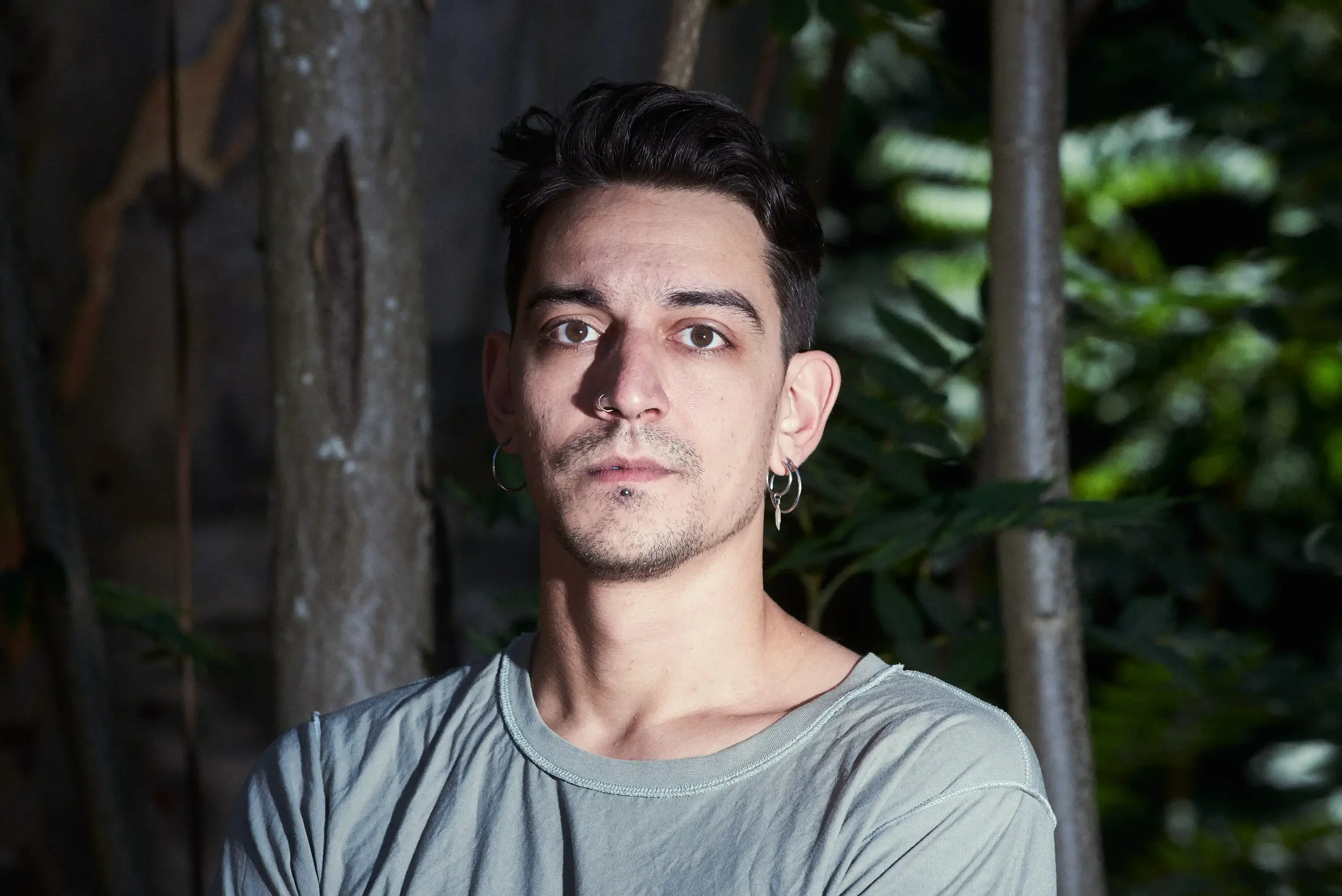 A young man with dark hair, earrings, and a nose ring standing outdoors in front of a tree and green foliage, looking directly at the camera.