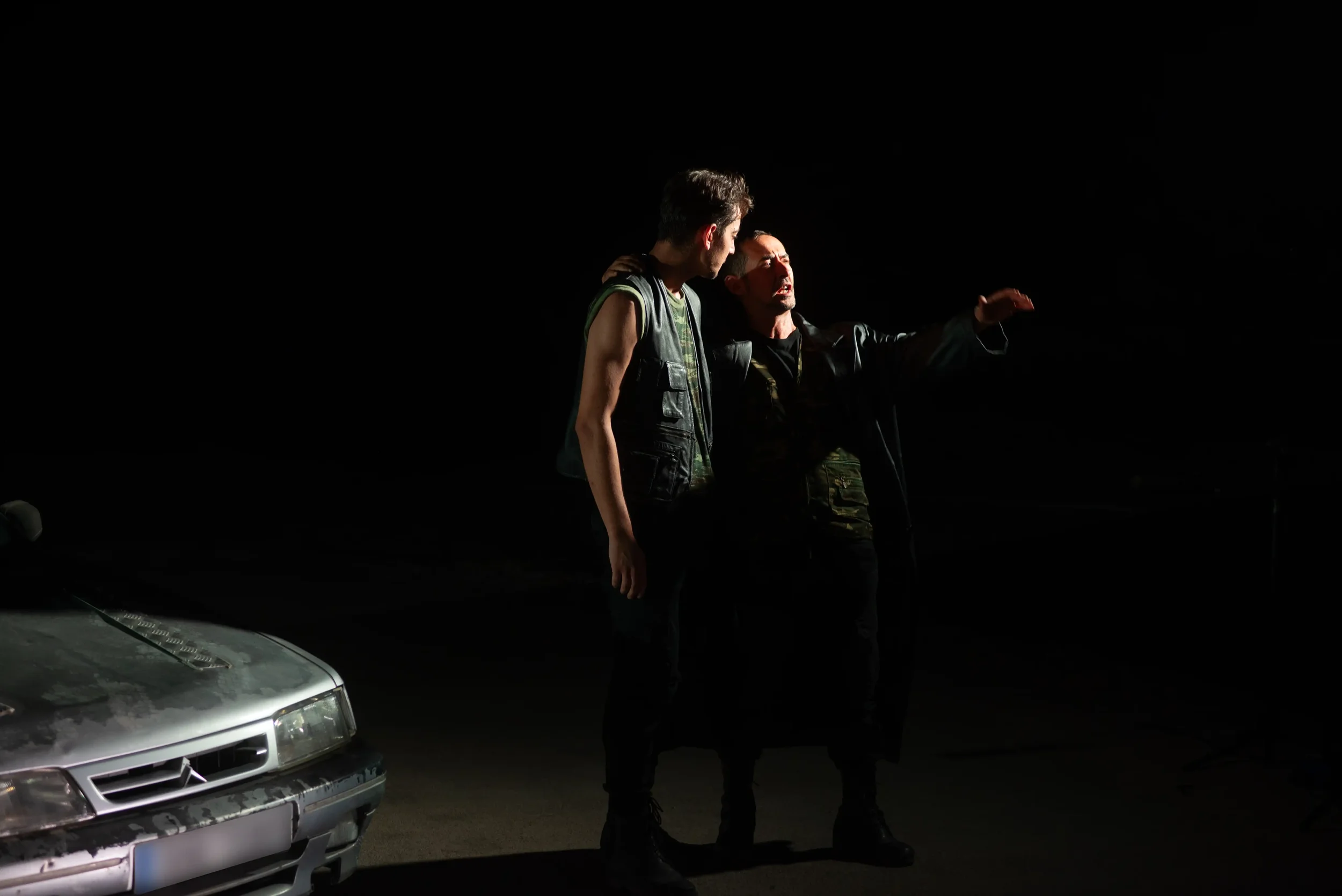 Two men talking on a dark street at night, one with a shoulder bag, one gesturing with his hand, and a rusty car in the foreground.