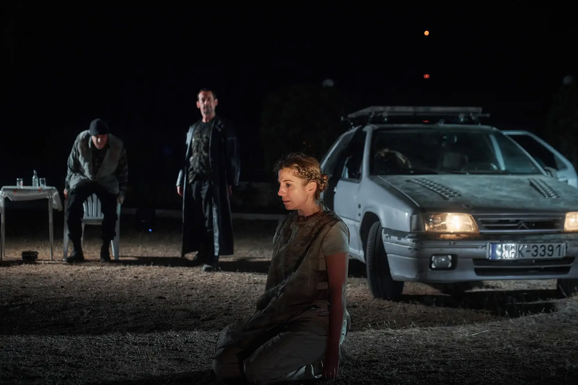 A woman kneeling on the ground at night, with three men standing behind her, near a white car.