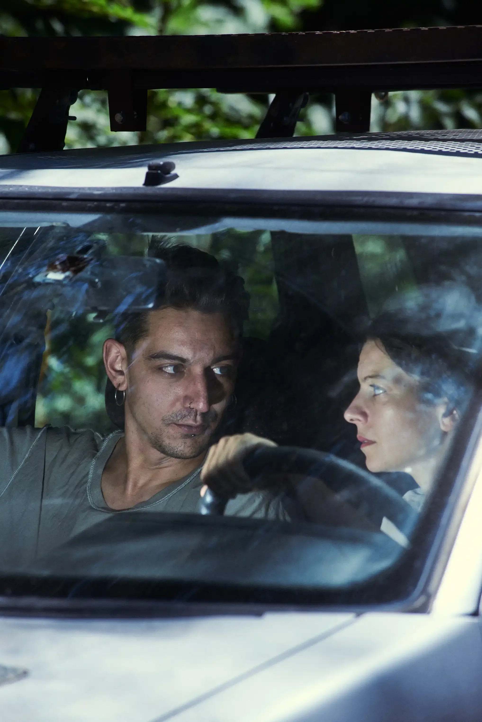 A man and a woman sitting inside a car, engaging in a conversation through the windshield. The woman is in the driver's seat and the man is in the passenger seat, both looking at each other.